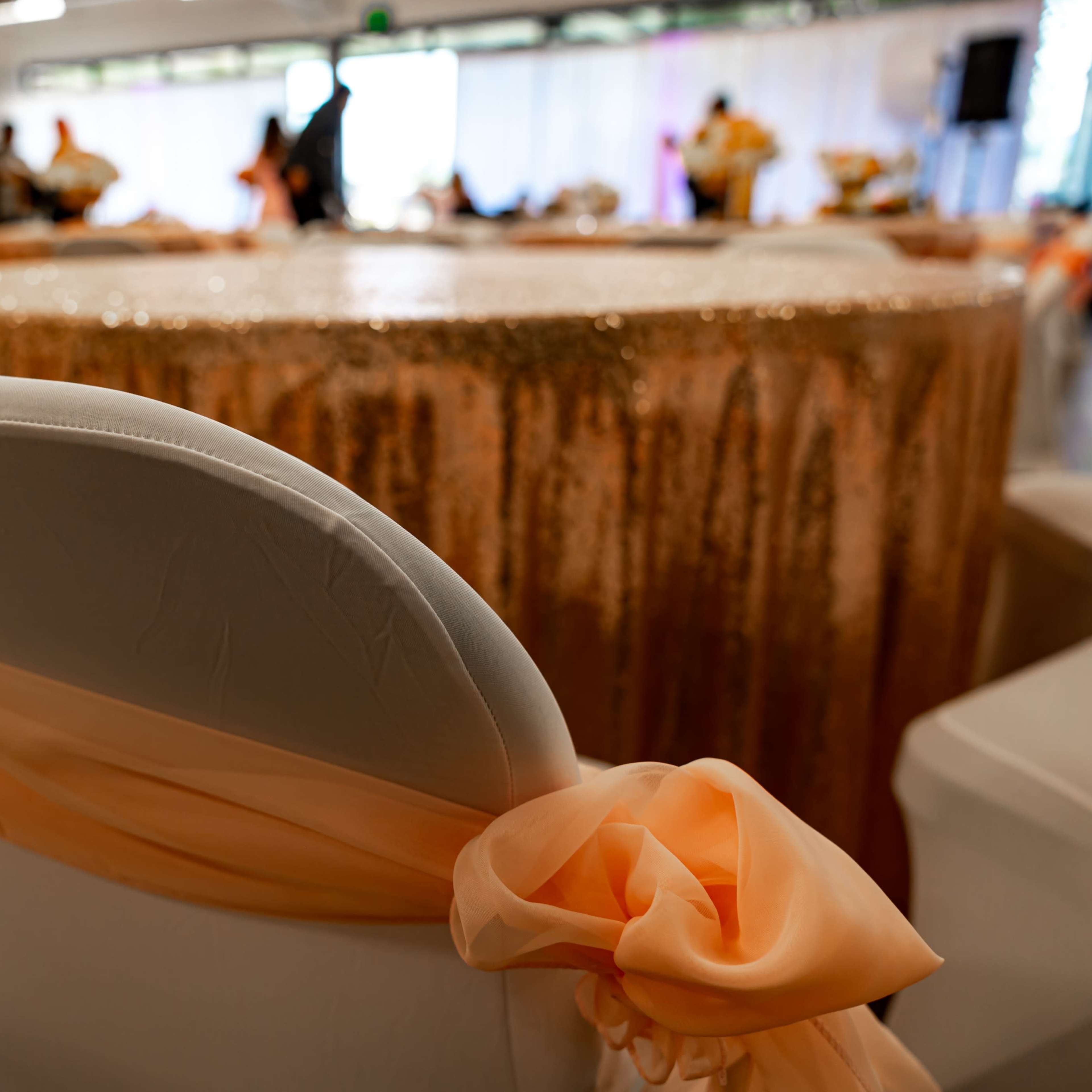 A decorated banquet hall features a table with a sequin tablecloth and chairs adorned with orange sashes.