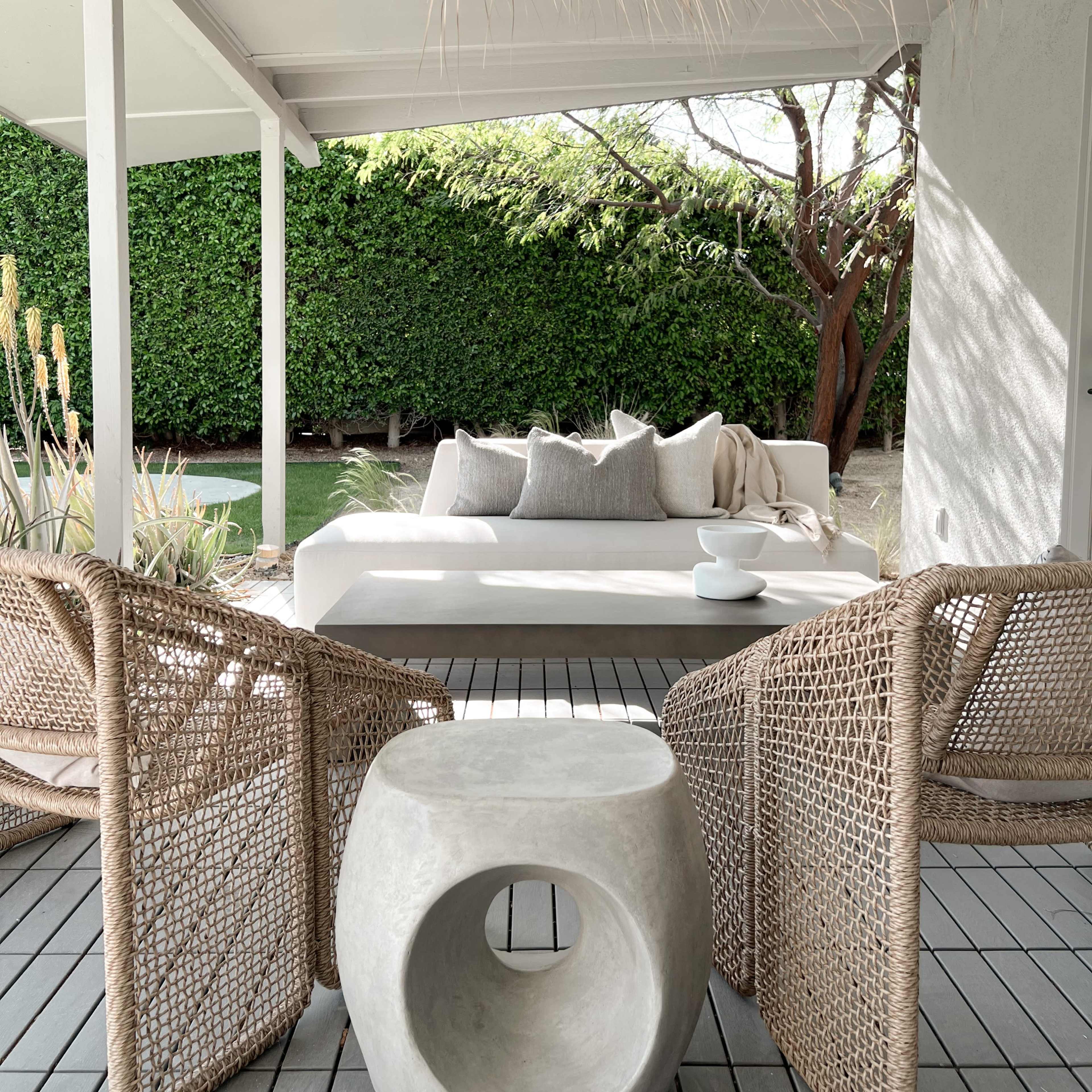 A modern outdoor seating area features a white sofa with cushions, two woven chairs, and a circular concrete side table against a backdrop of green foliage.