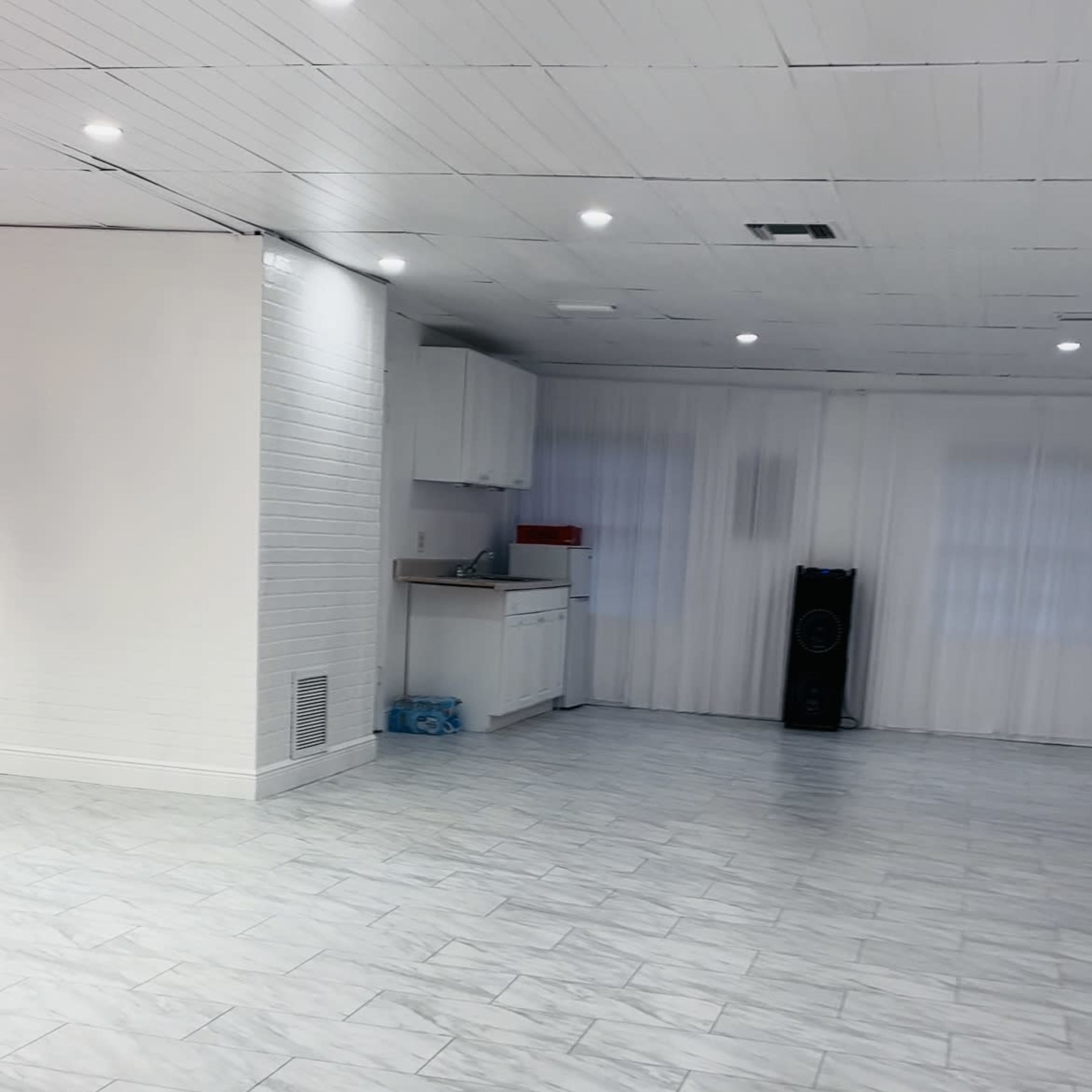 A large, empty room with white tiled flooring, a kitchenette in the corner, and large windows covered with sheer curtains.