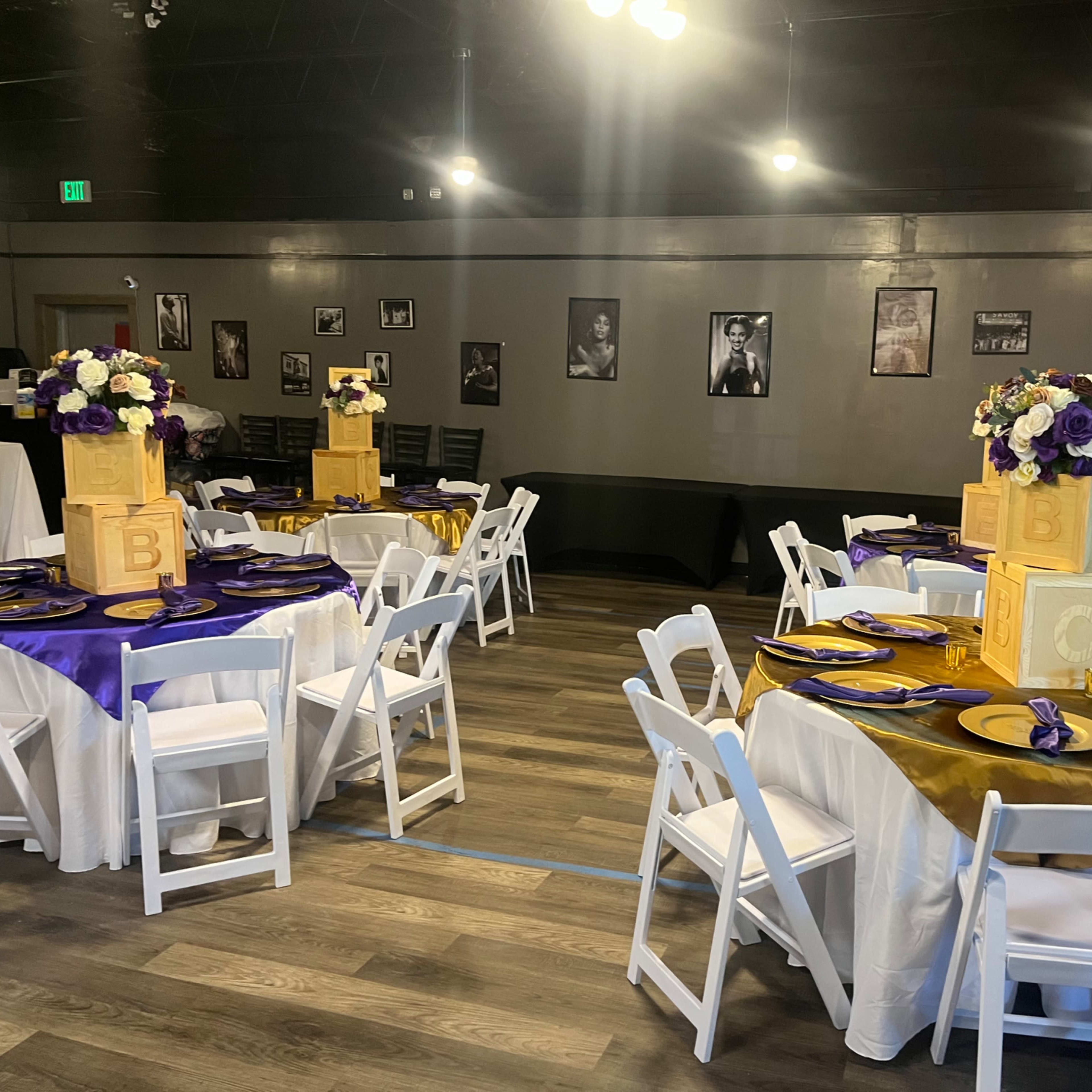 A banquet hall with several round tables decorated with white tablecloths, gold and purple accents, and floral centerpieces, surrounded by chairs and framed photographs on the walls.