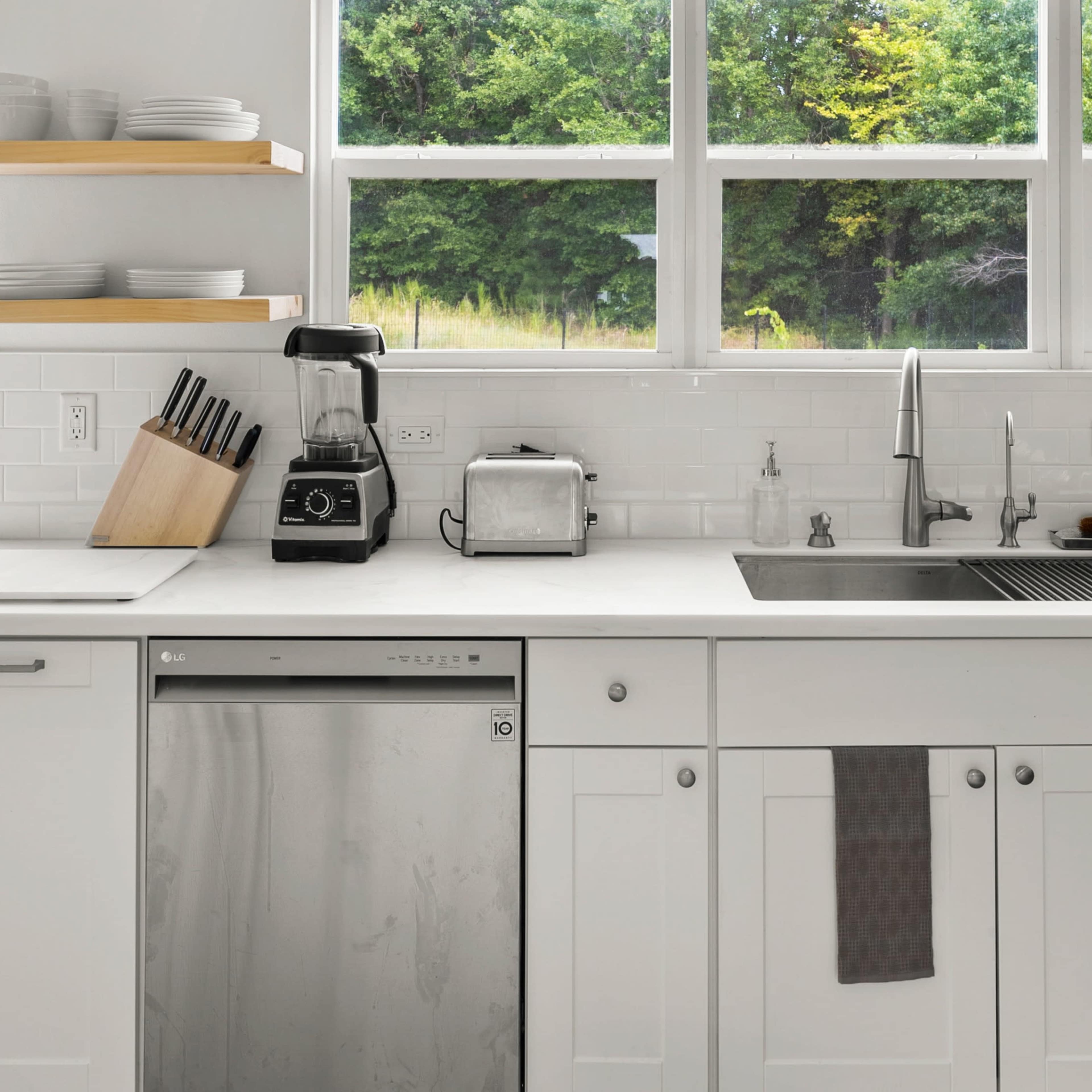 A modern kitchen features white cabinetry, a stainless steel dishwasher, and a counter with a blender and toaster beside large windows overlooking greenery.