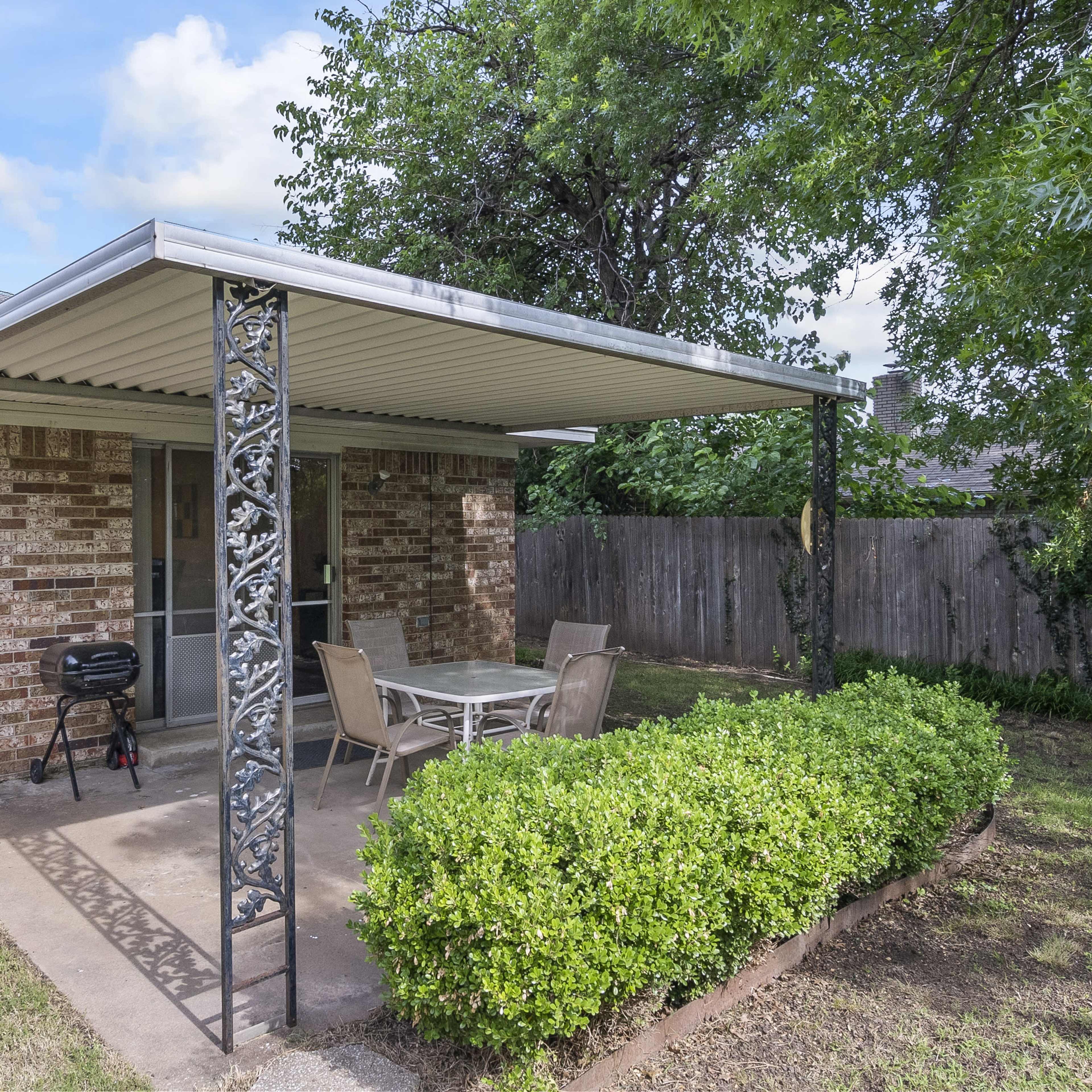 A patio area features a metal canopy with a table and chairs, bordered by a small hedge and a grill nearby, set against a brick wall and landscaped yard.