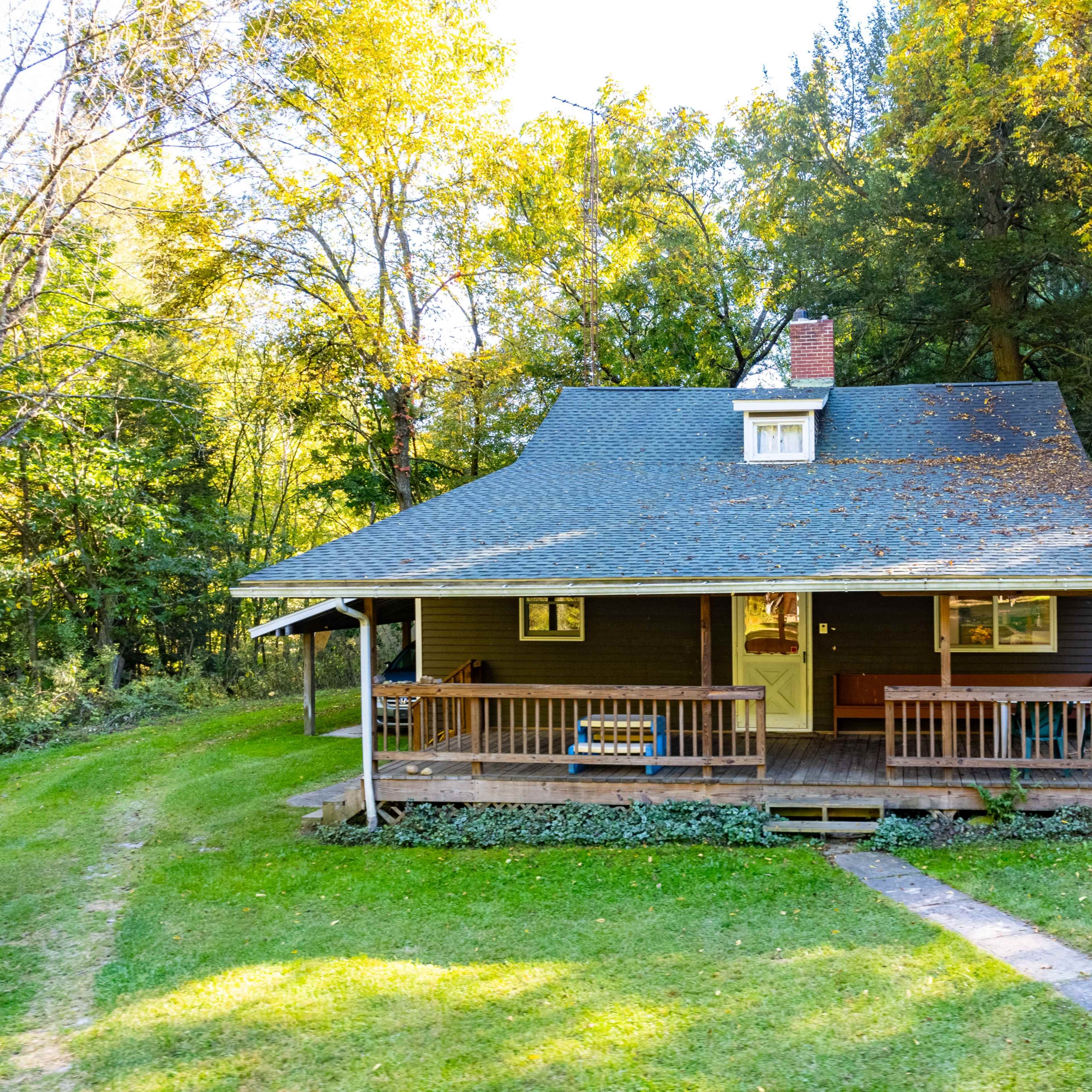 A rustic house with a sloped roof and a front porch is surrounded by trees and a grassy lawn.