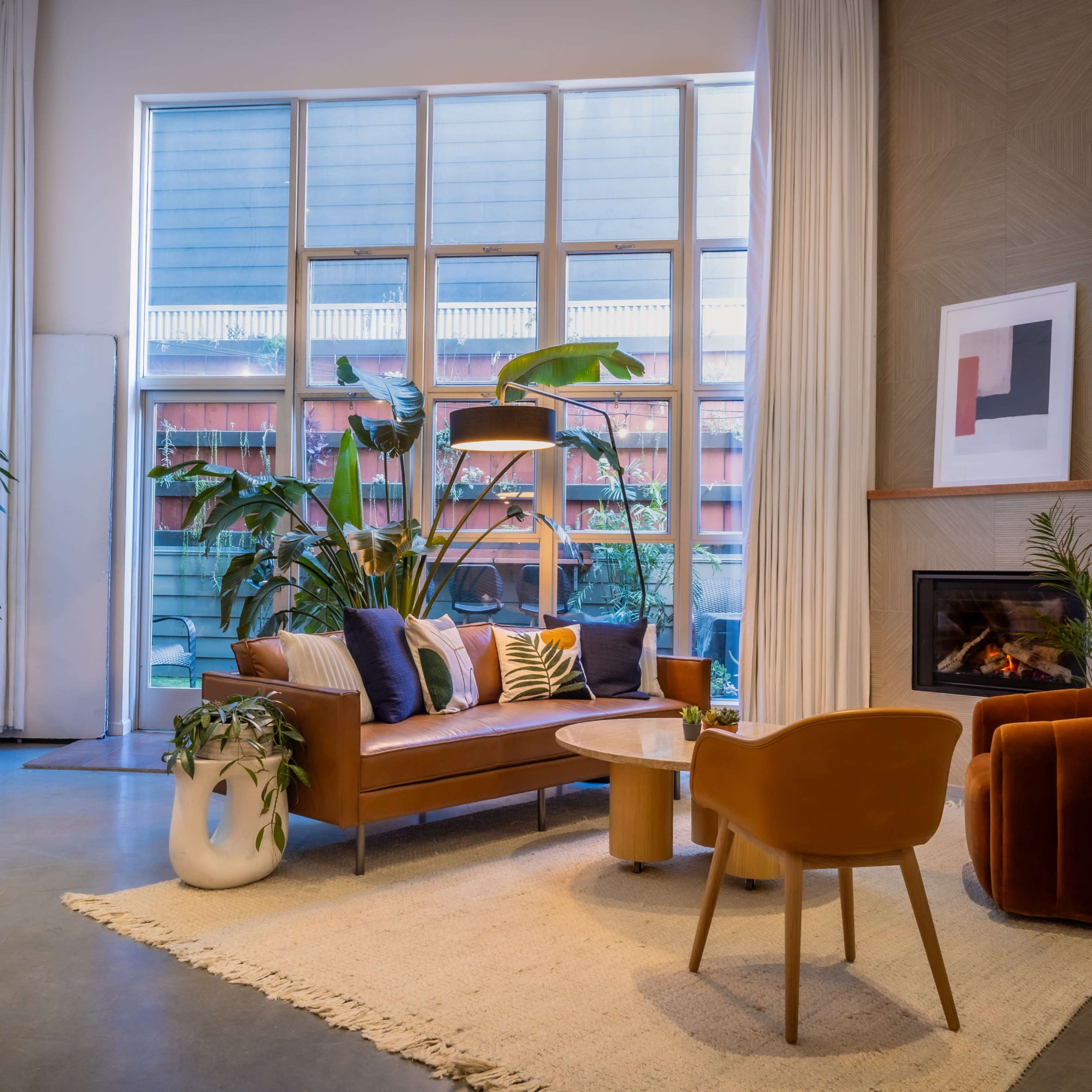 A modern living room with a brown leather sofa, a round wooden coffee table, and large windows filled with greenery.