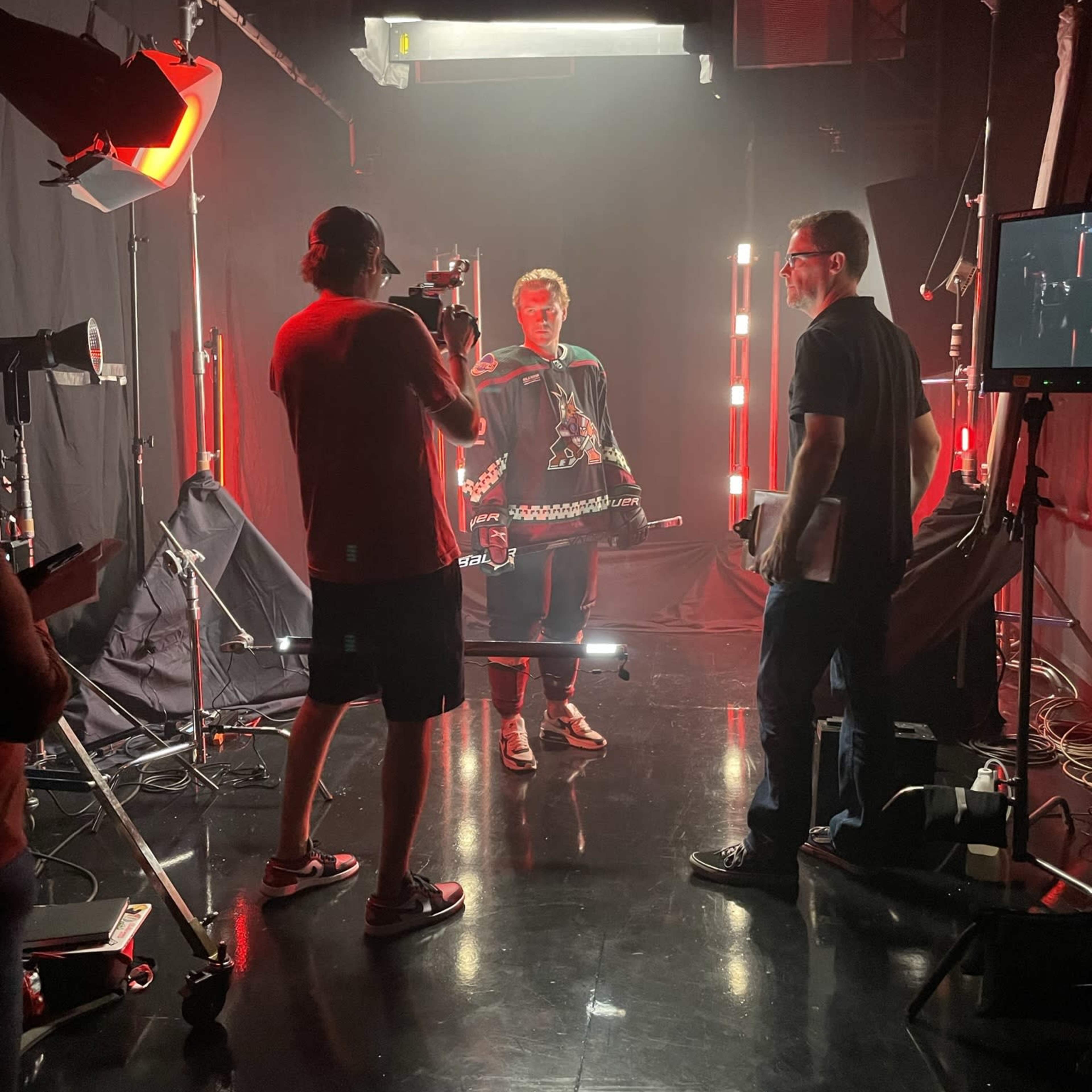A person in a dark outfit stands in a studio surrounded by bright red lights while crew members operate cameras and equipment.