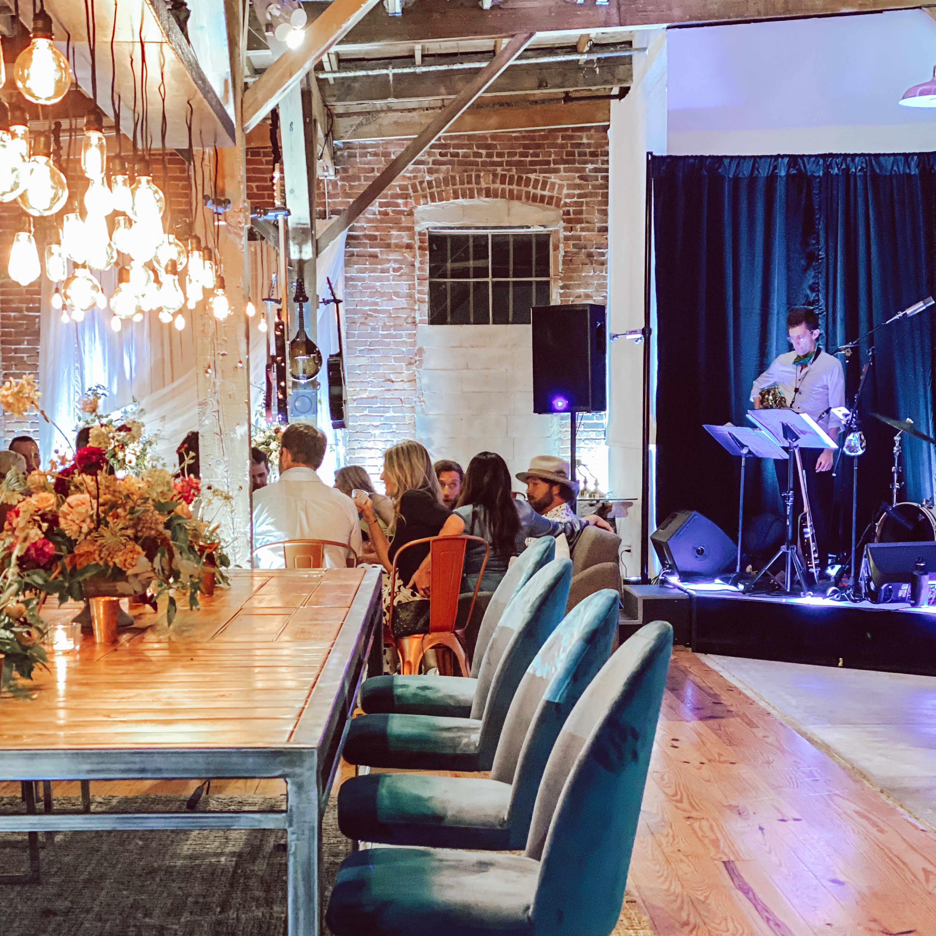 A spacious event venue with a long dining table, industrial-style lighting, and a small stage where a musician performs.