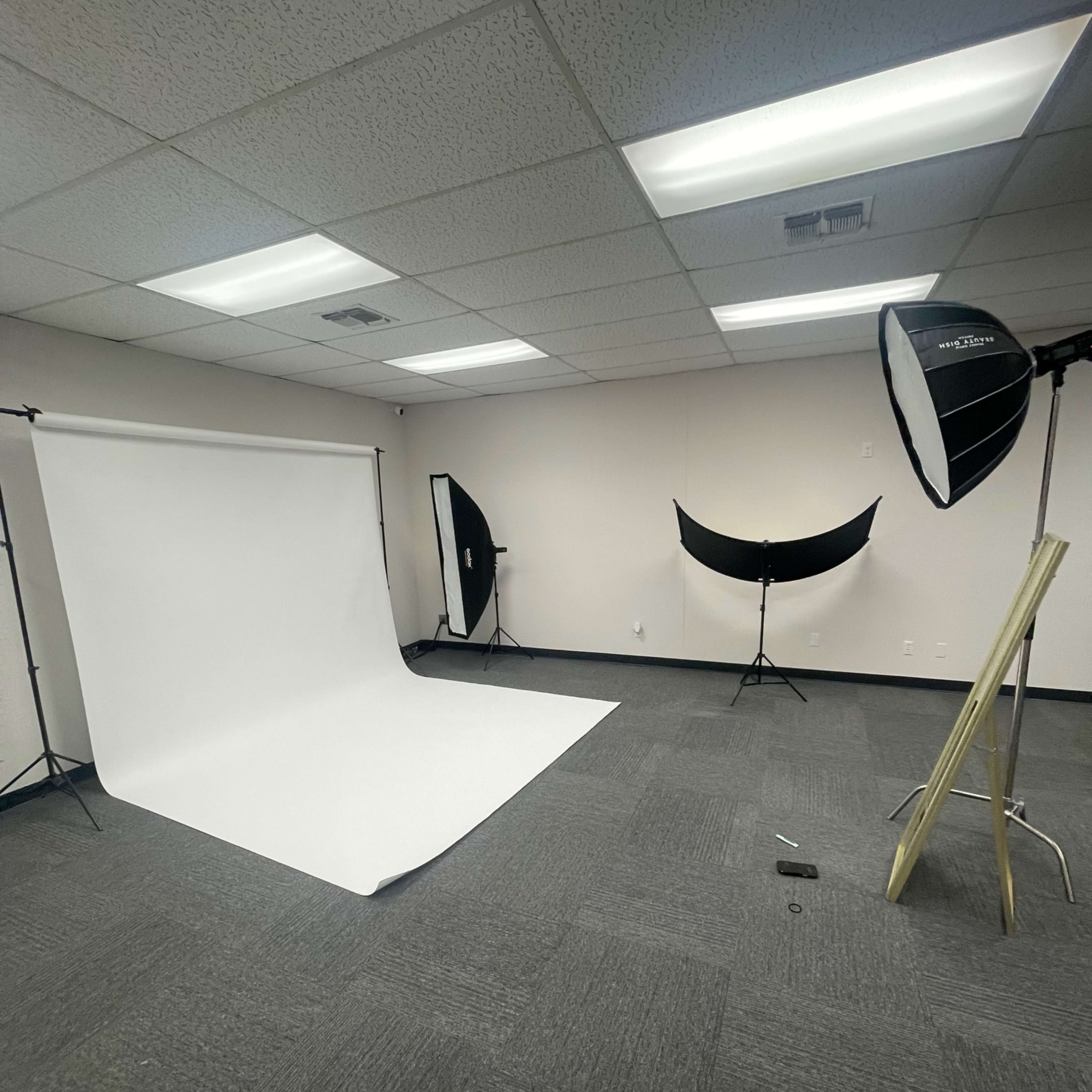 A photography studio with a white backdrop, two light softboxes, and a reflector set up in an empty room.