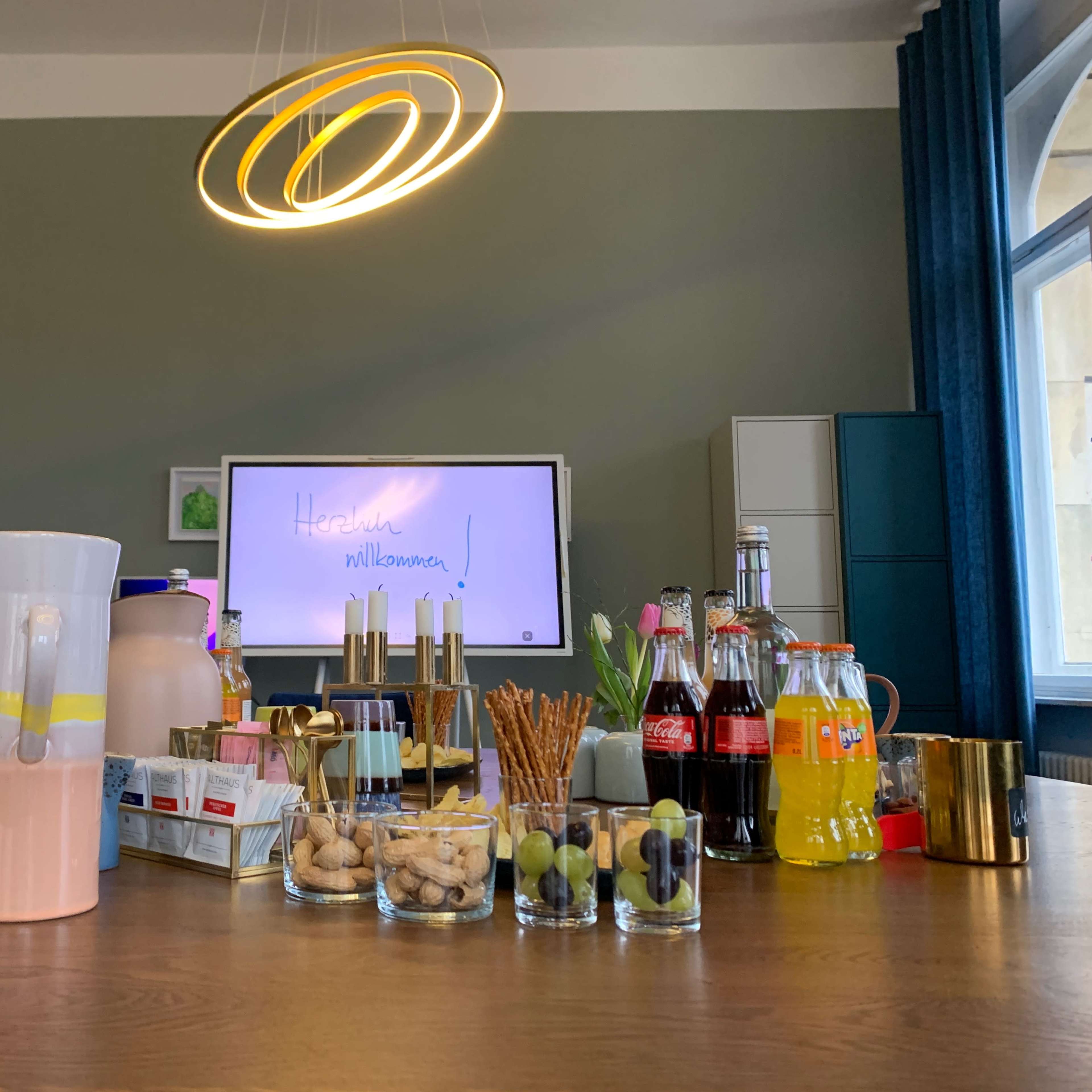 A table is set with various beverages, snacks, and flowers in a room featuring a large screen and modern decor.
