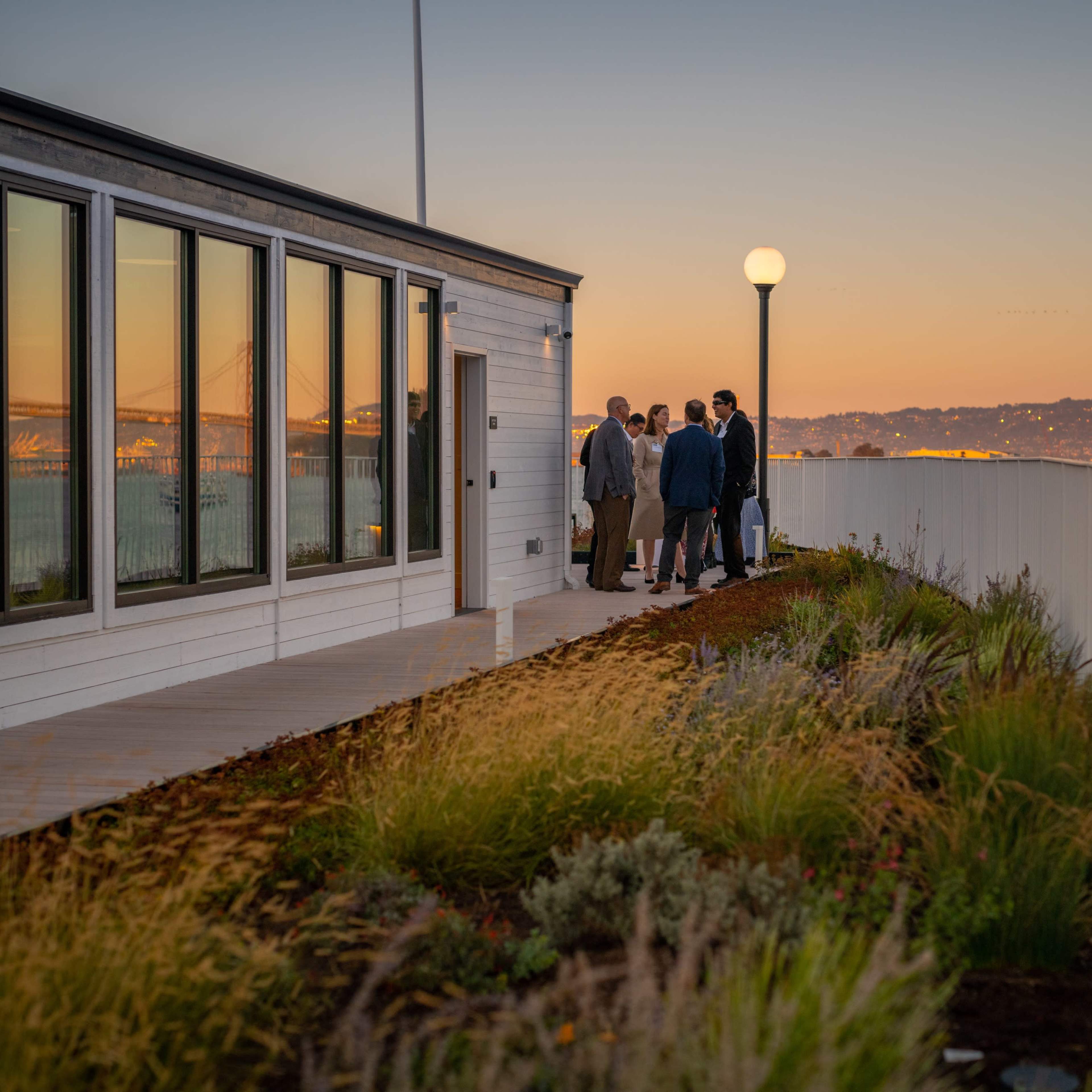 A group of people stands on a patio near a white building, overlooking a waterfront with a distant bridge at sunset.
