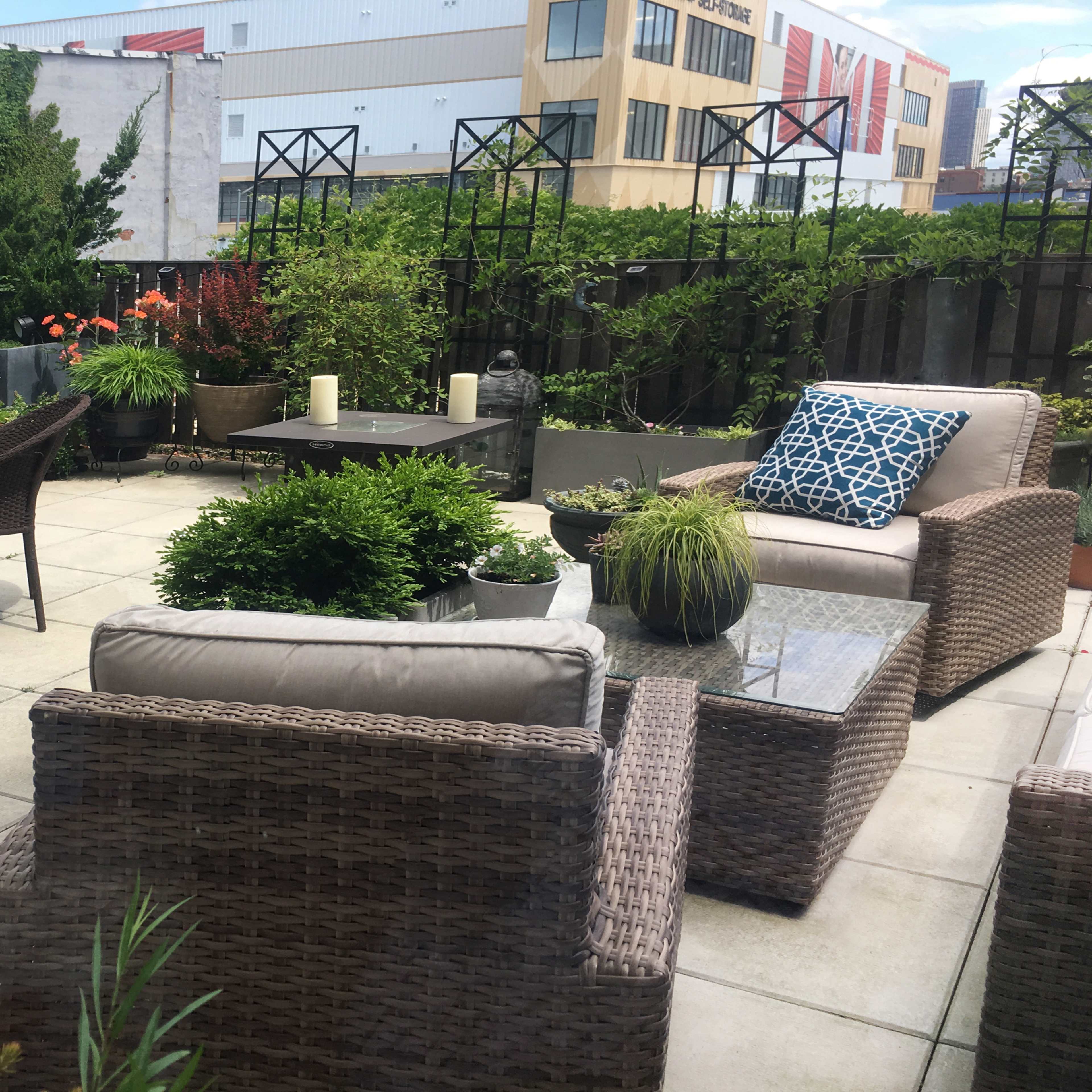 A patio area with wicker furniture, potted plants, and a dining table surrounded by greenery and urban buildings in the background.