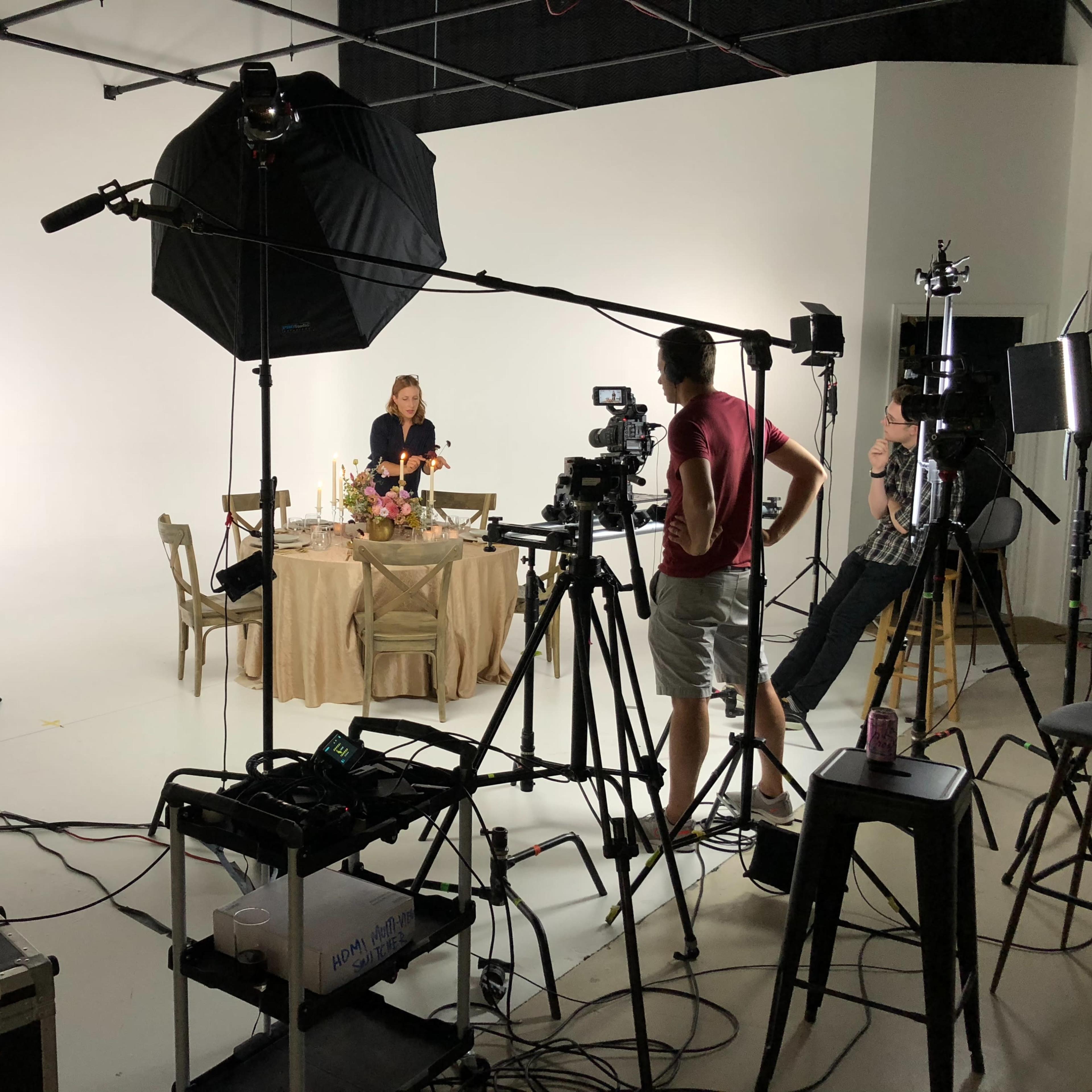 A film crew sets up equipment around a table decorated with flowers in a well-lit studio.