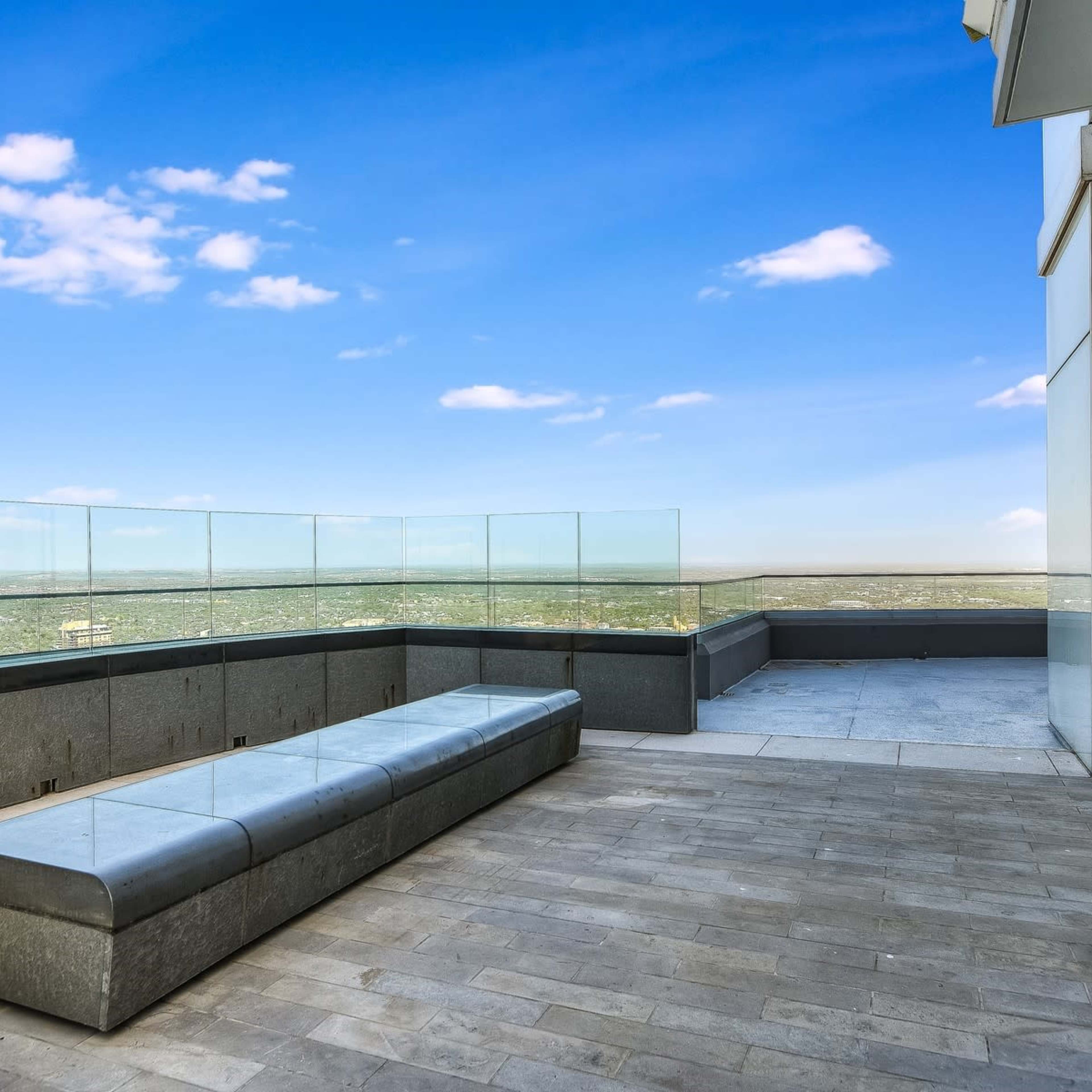 A spacious rooftop terrace features a stone bench and glass railing overlooking a vast landscape under a blue sky.