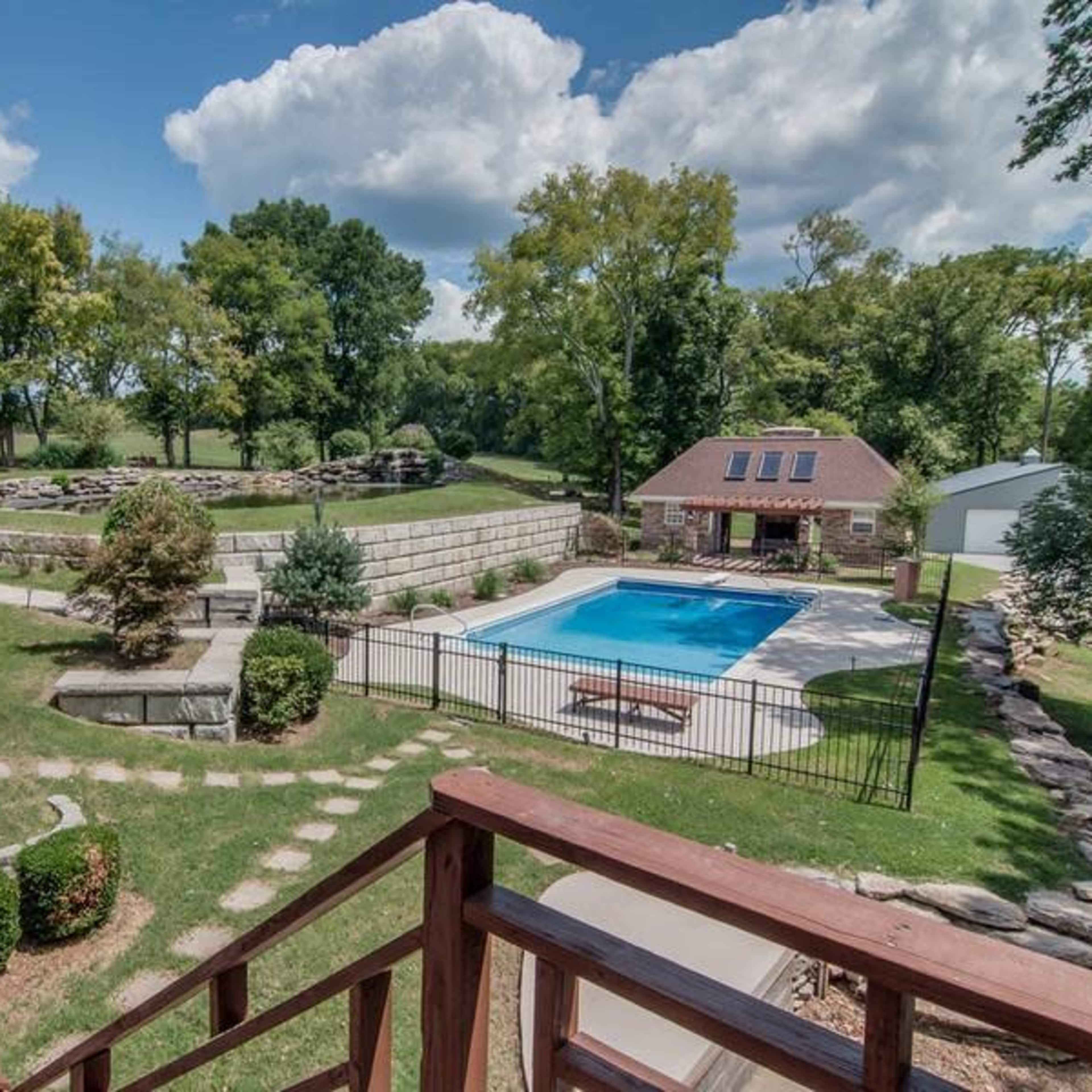 A landscaped backyard featuring a swimming pool, surrounding trees, a stone wall, and a small building with solar panels.