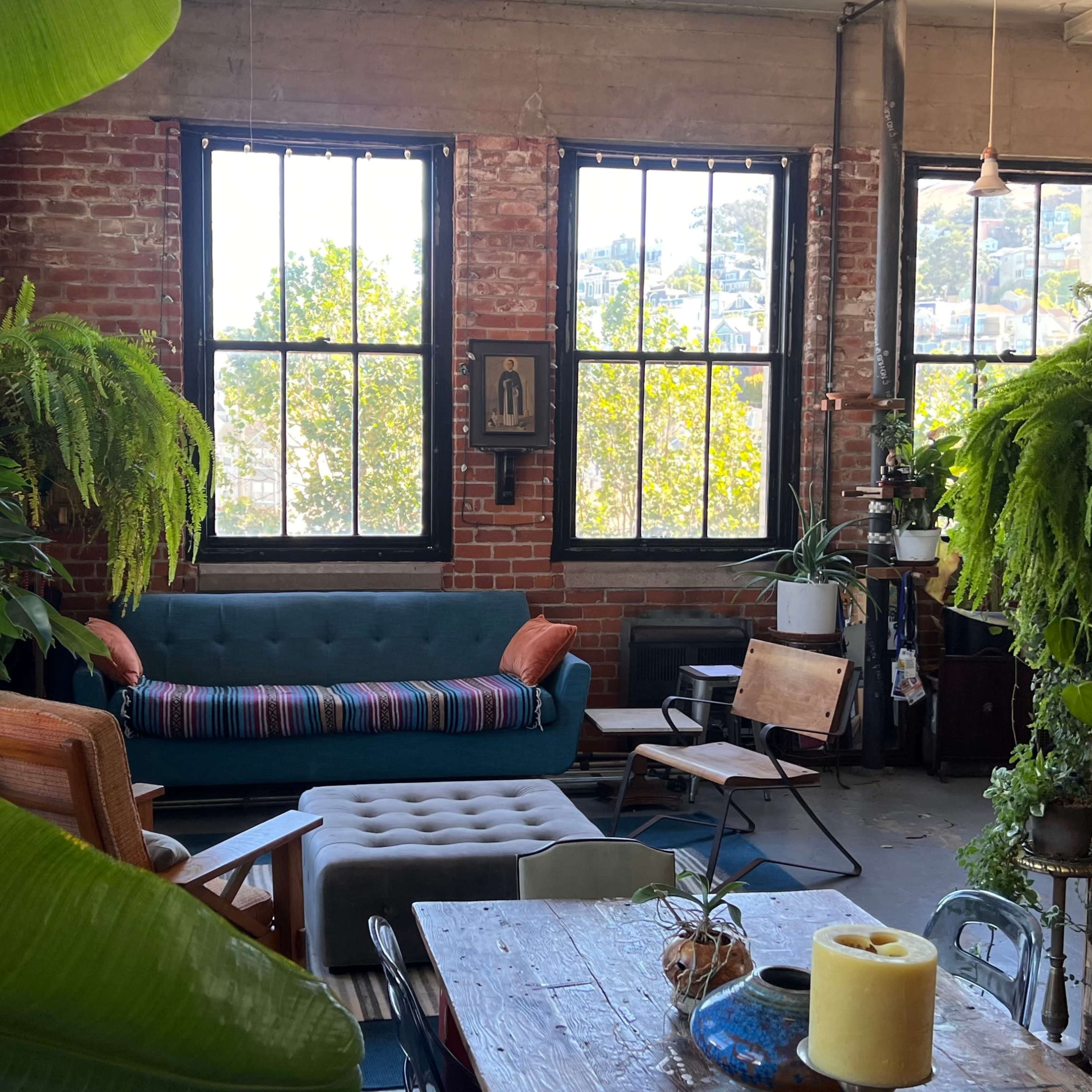 A cozy living space with large windows, a green couch, a wooden table, and various indoor plants.