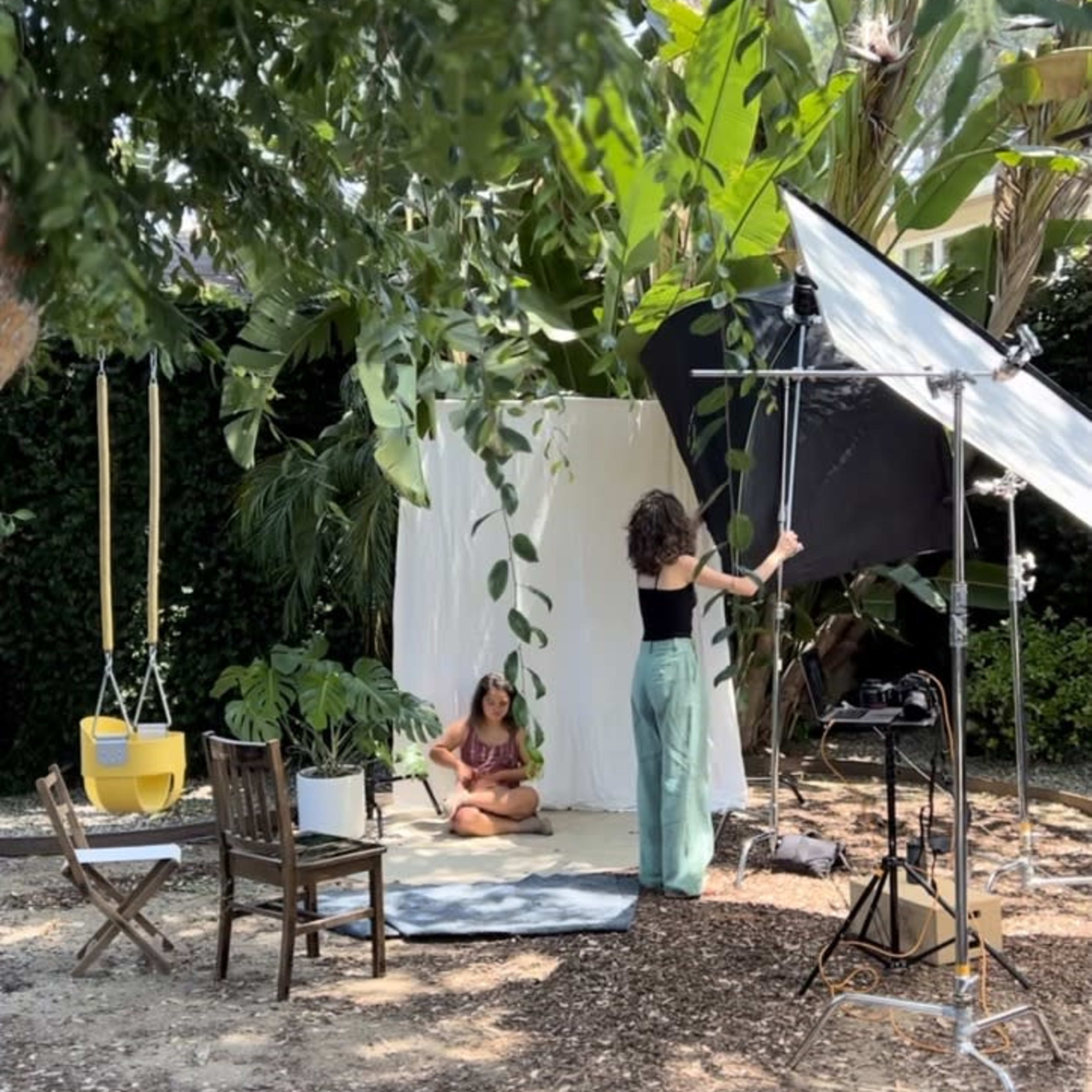 A woman adjusts lighting equipment while another sits on a mat in a garden setting surrounded by tropical plants.