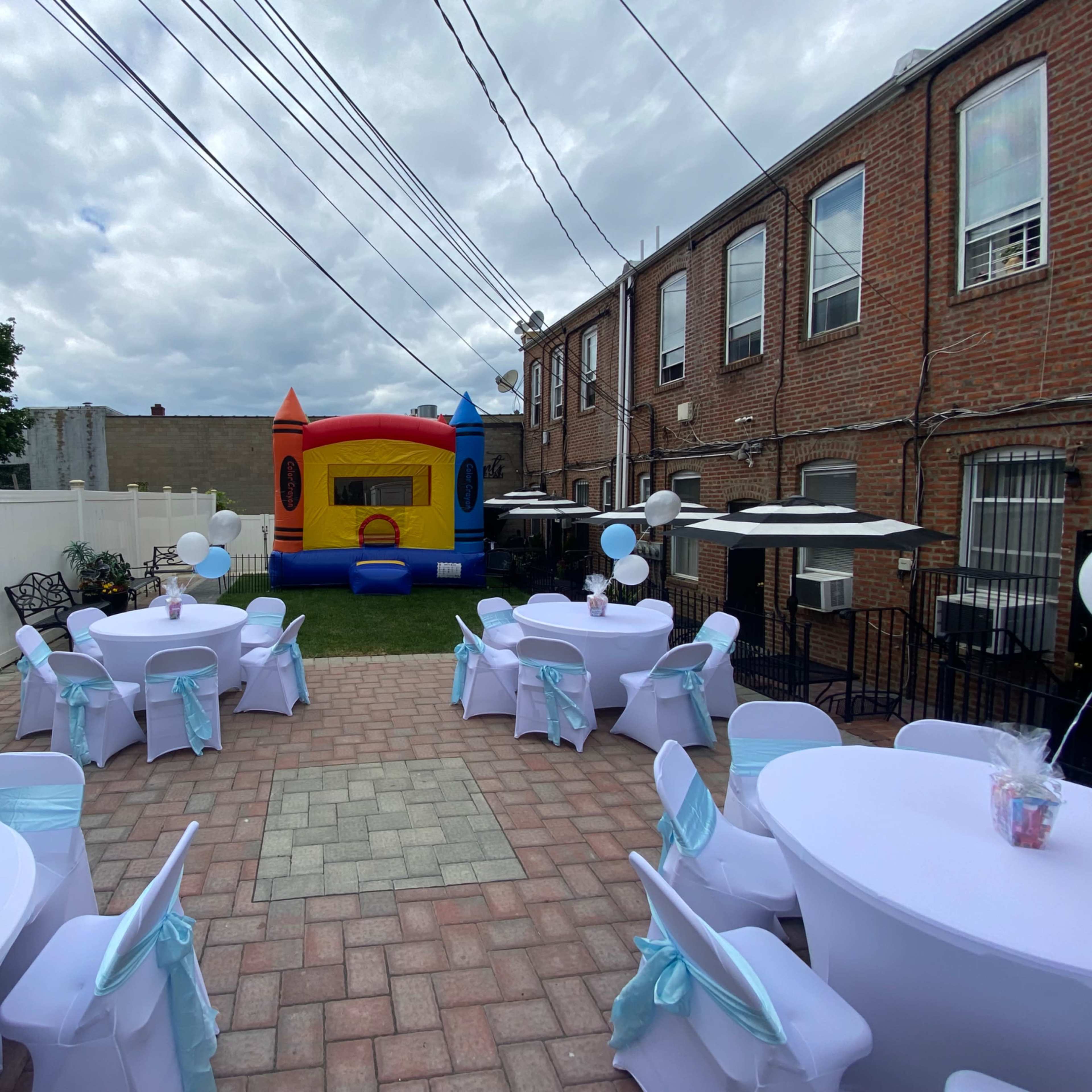 A backyard party setup features round tables with white tablecloths and light blue chair sashes, alongside a colorful inflatable bouncing castle.