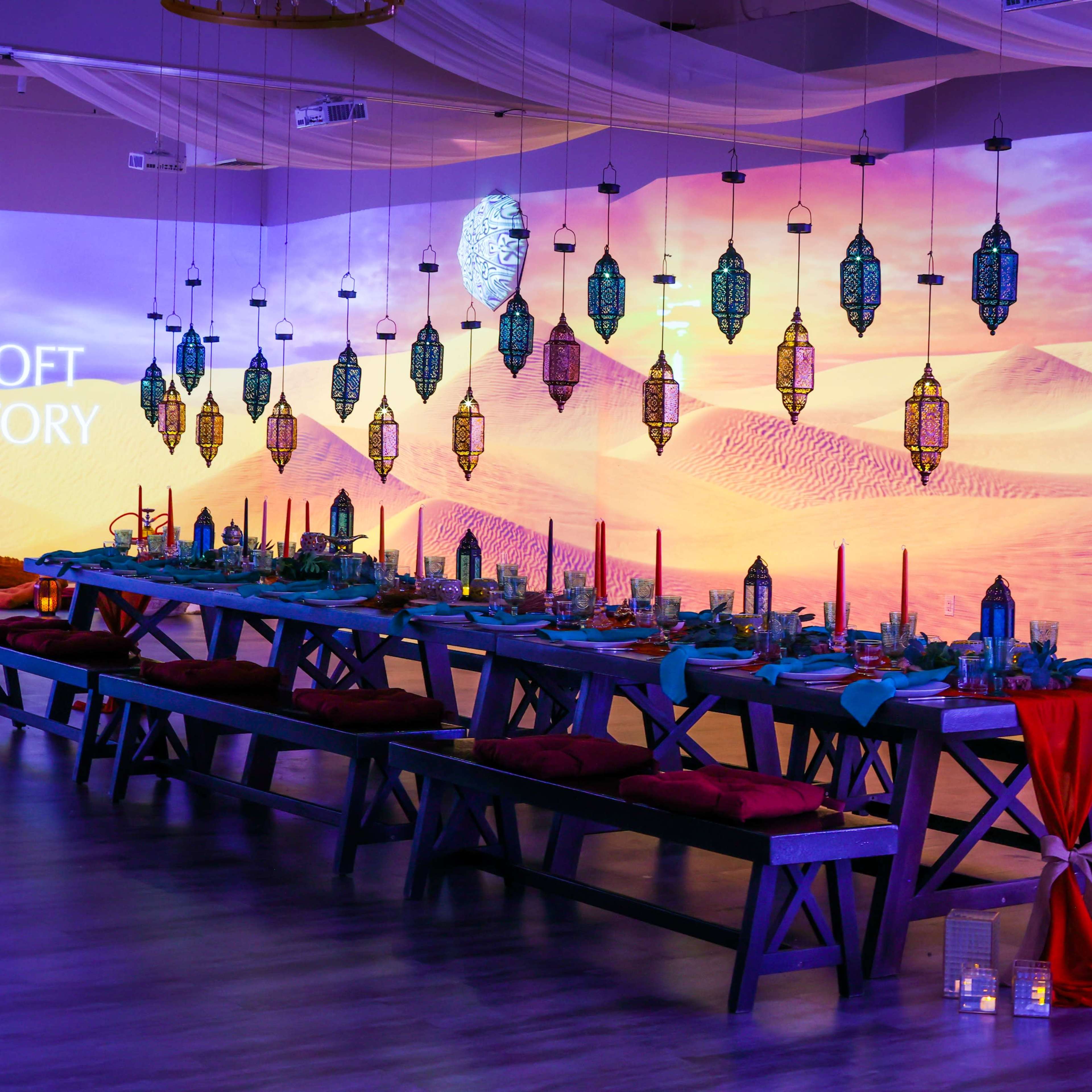 The scene features a long dining table adorned with decorative lanterns and cushions, set against a backdrop of an illuminated desert landscape.