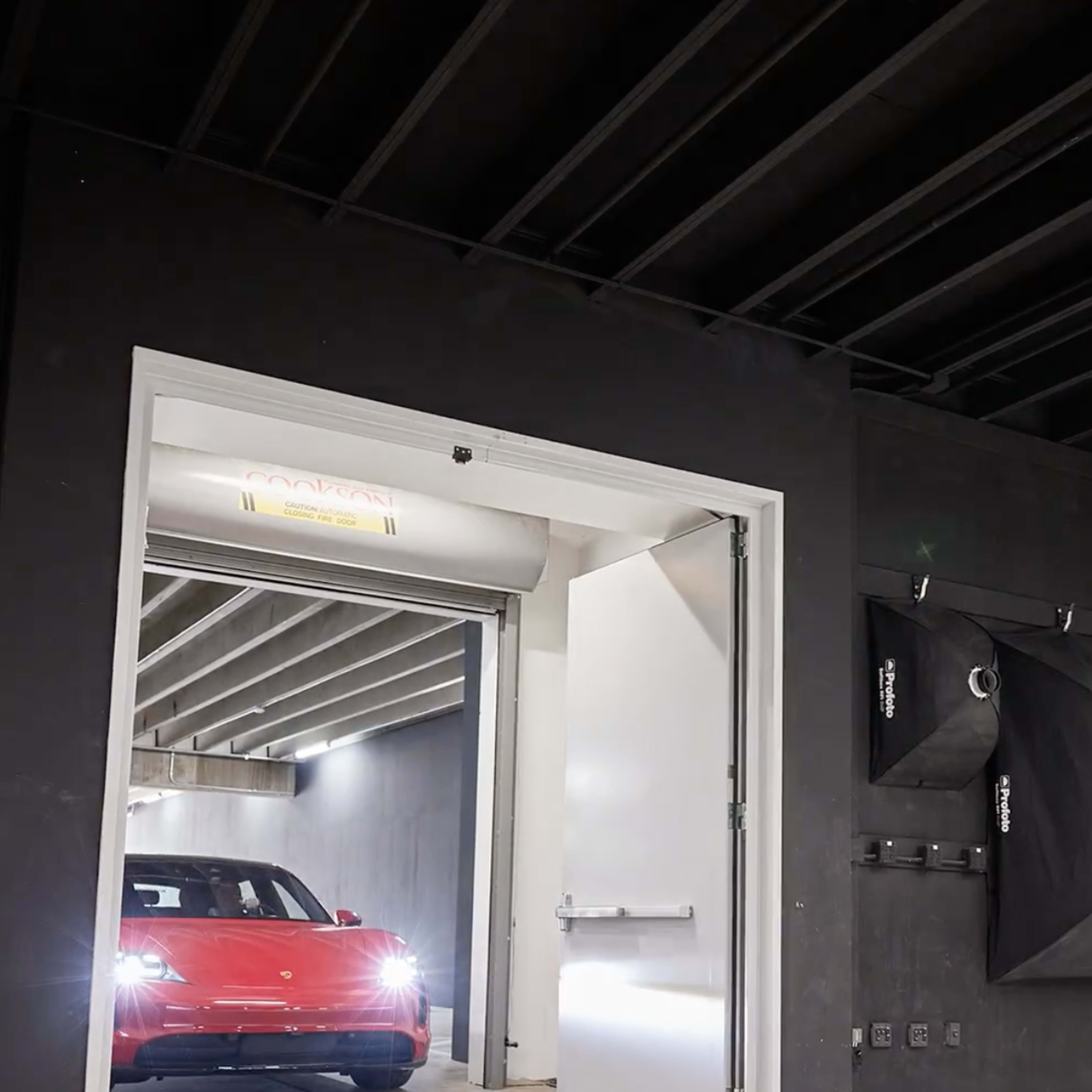 A red car is parked at the entrance of a dimly lit garage with an open door and a black ceiling.