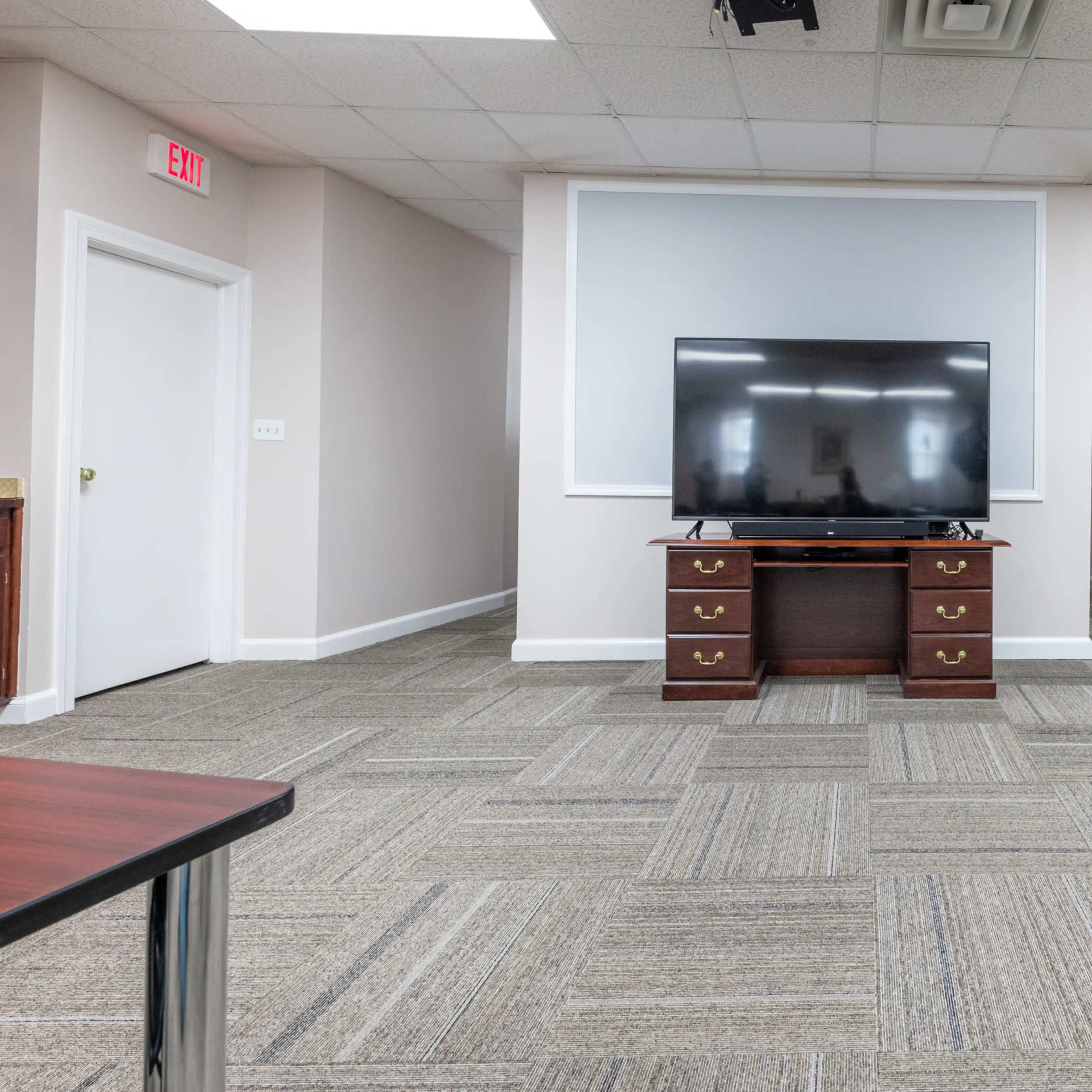 A spacious room features a wall-mounted television set on a dark wooden cabinet, with a gray carpet and light-colored walls.