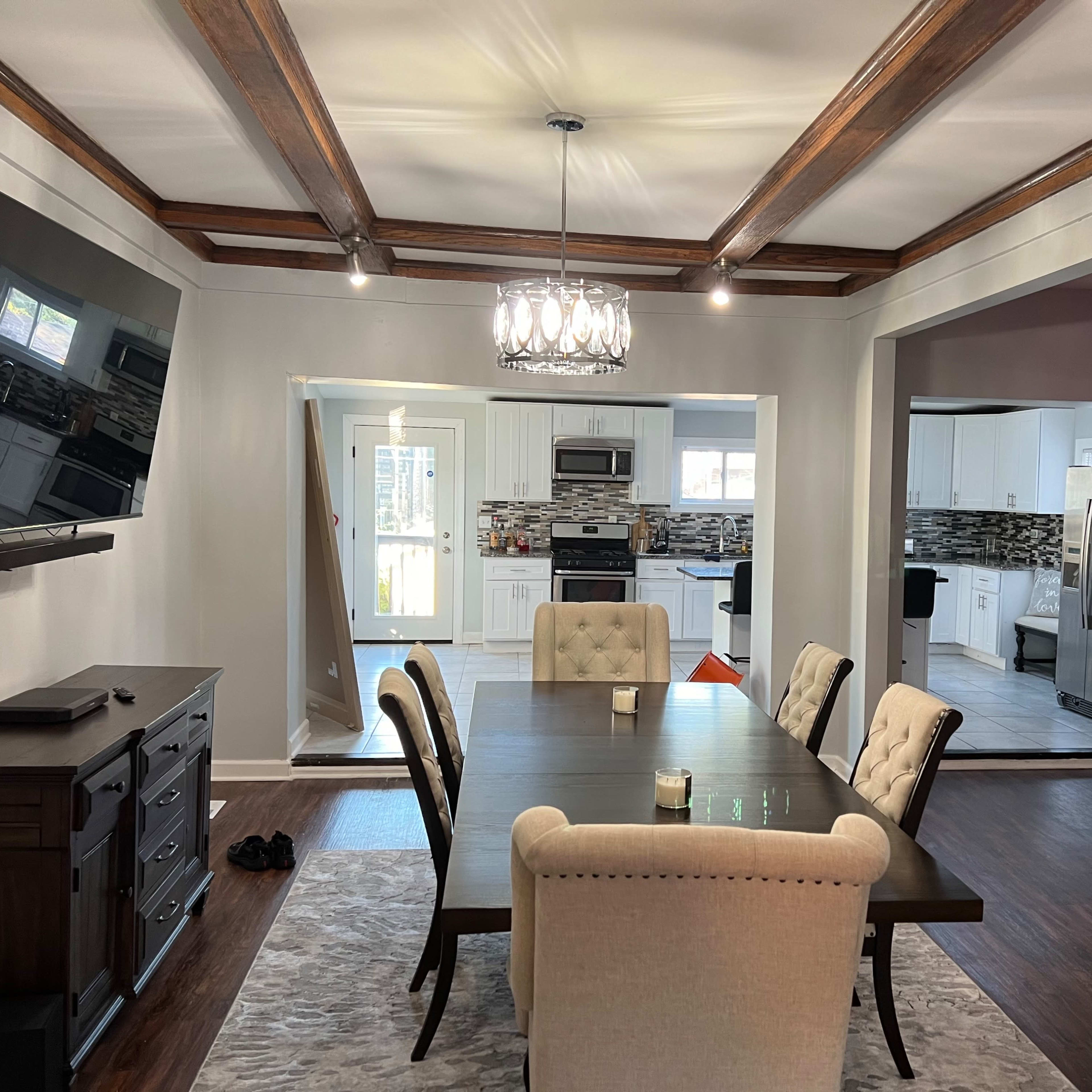 A dining area with a dark wooden table surrounded by upholstered chairs, a chandelier overhead, and an open view into a kitchen with modern appliances and decor.