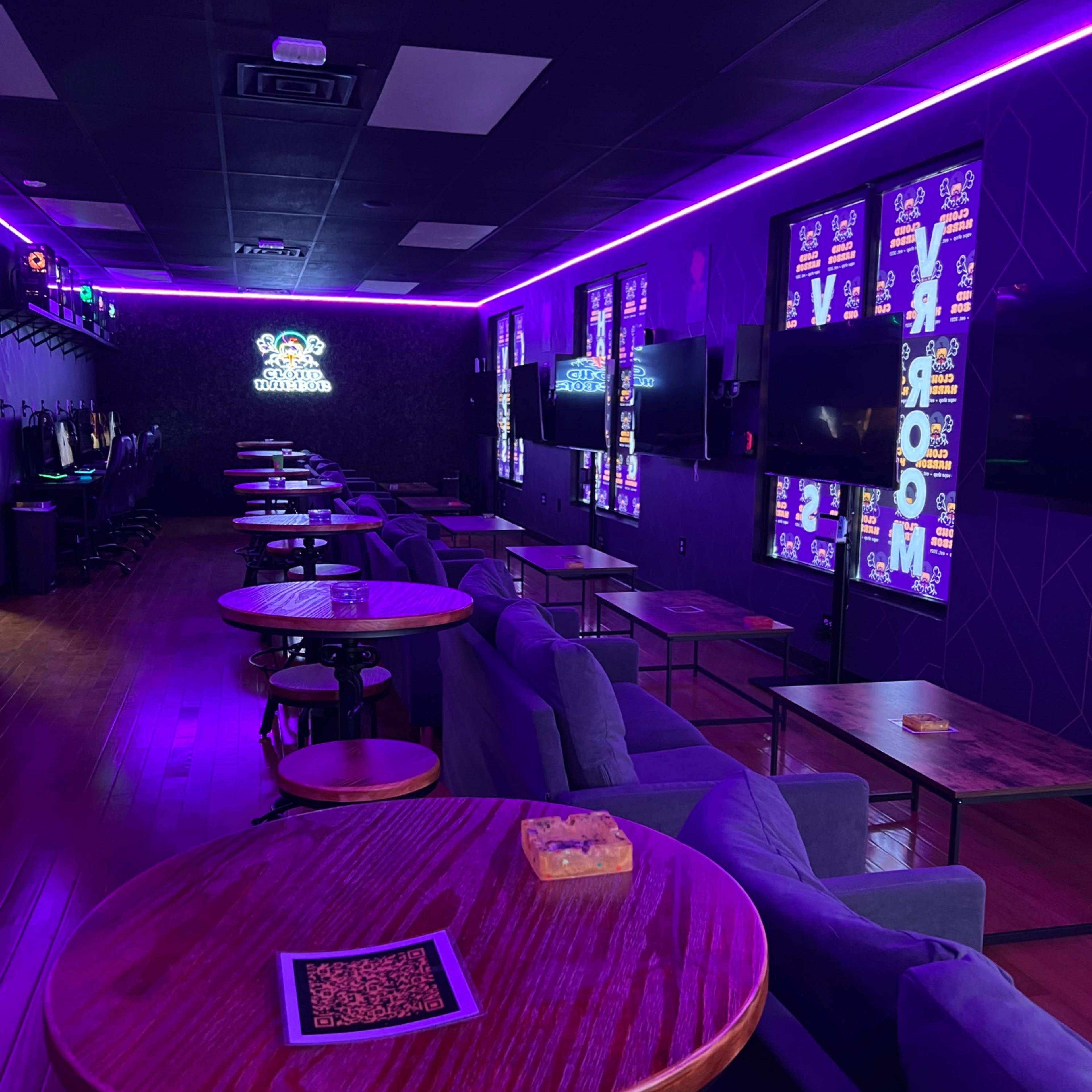 A dimly lit lounge area with purple neon lighting, featuring rows of tables and chairs along the walls equipped with multiple TVs.