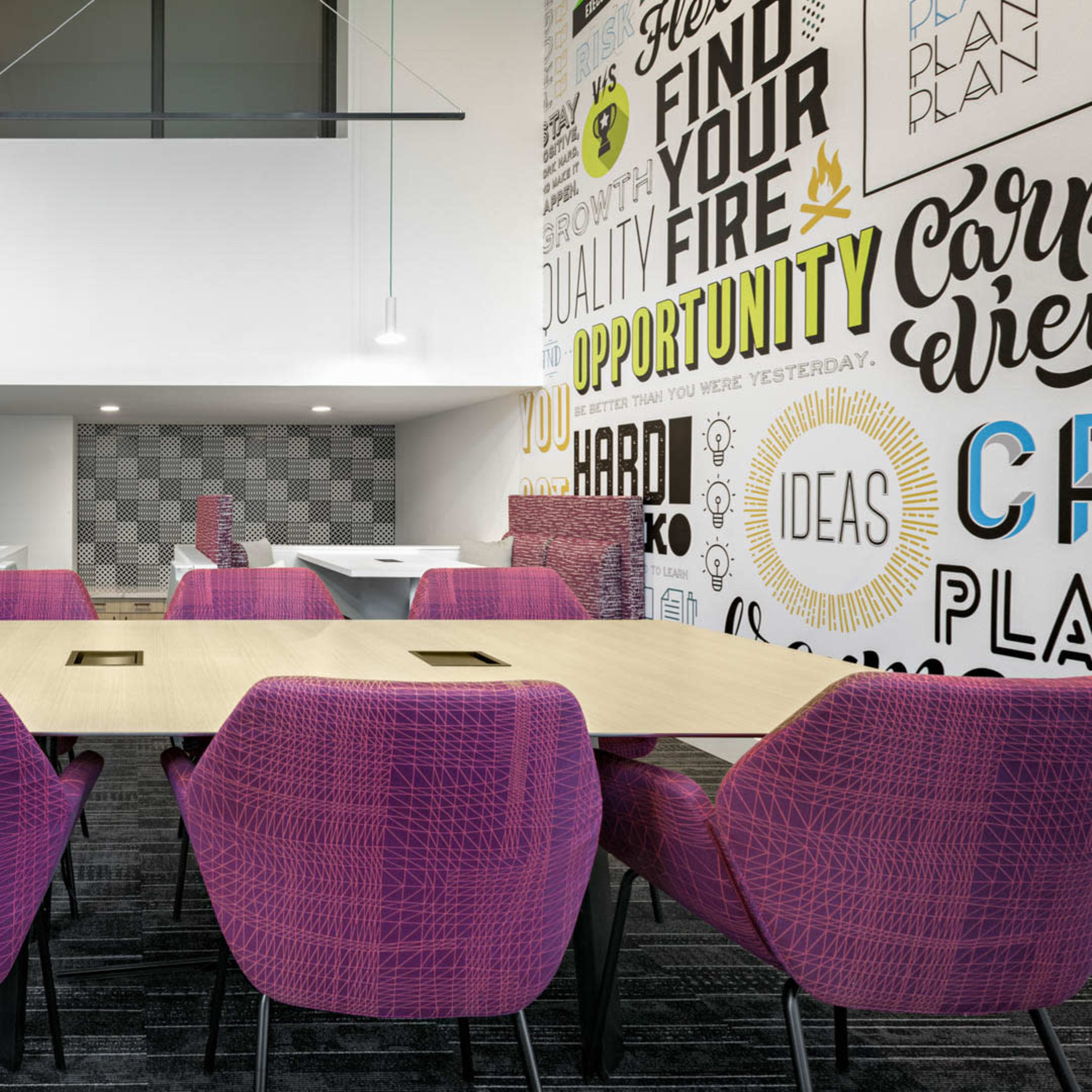A modern conference room with a large rectangular table surrounded by purple chairs, and a wall decorated with motivational phrases.