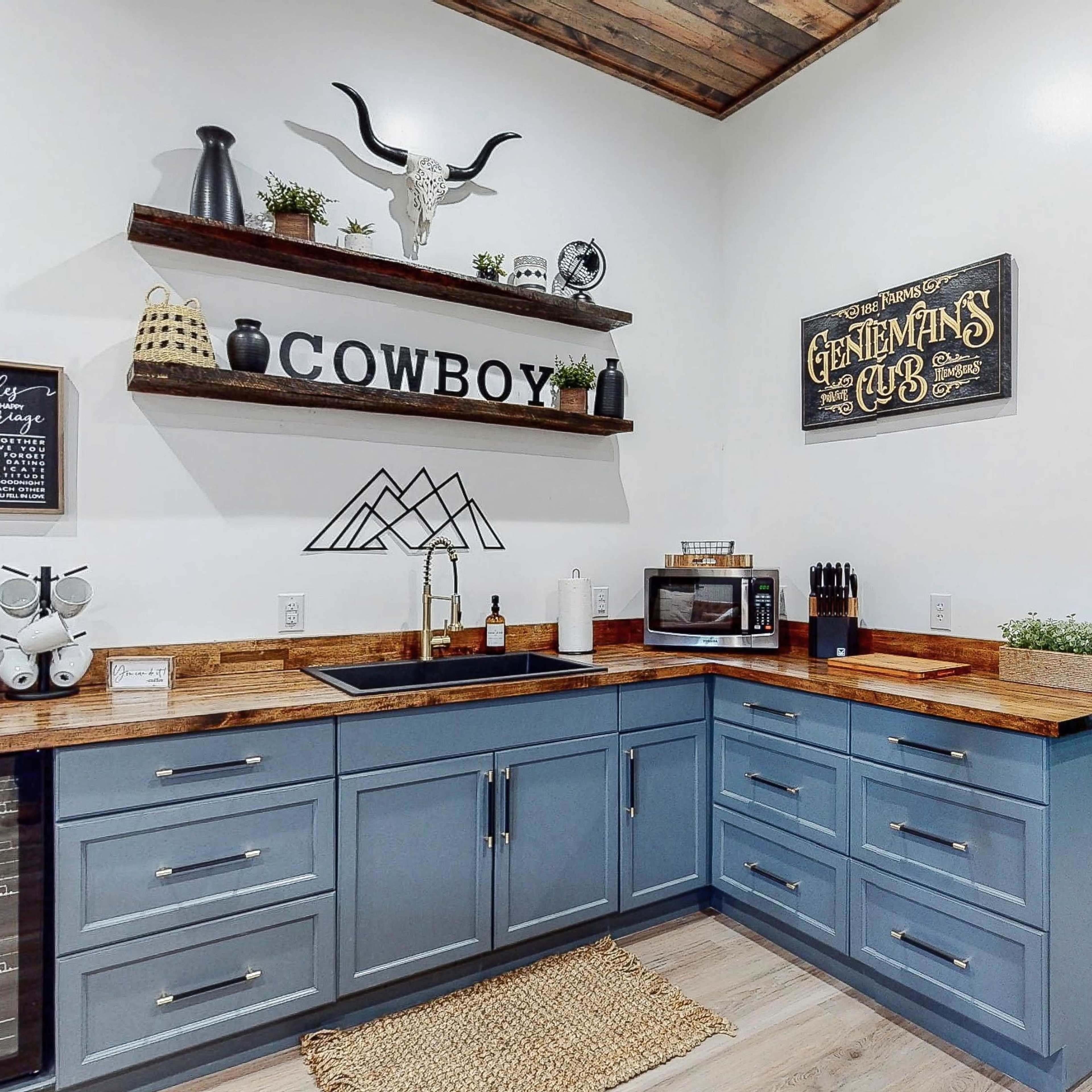 A kitchen corner with blue cabinetry, wooden countertops, a coffee machine, a microwave, and decorative shelves displaying a cowboy theme.