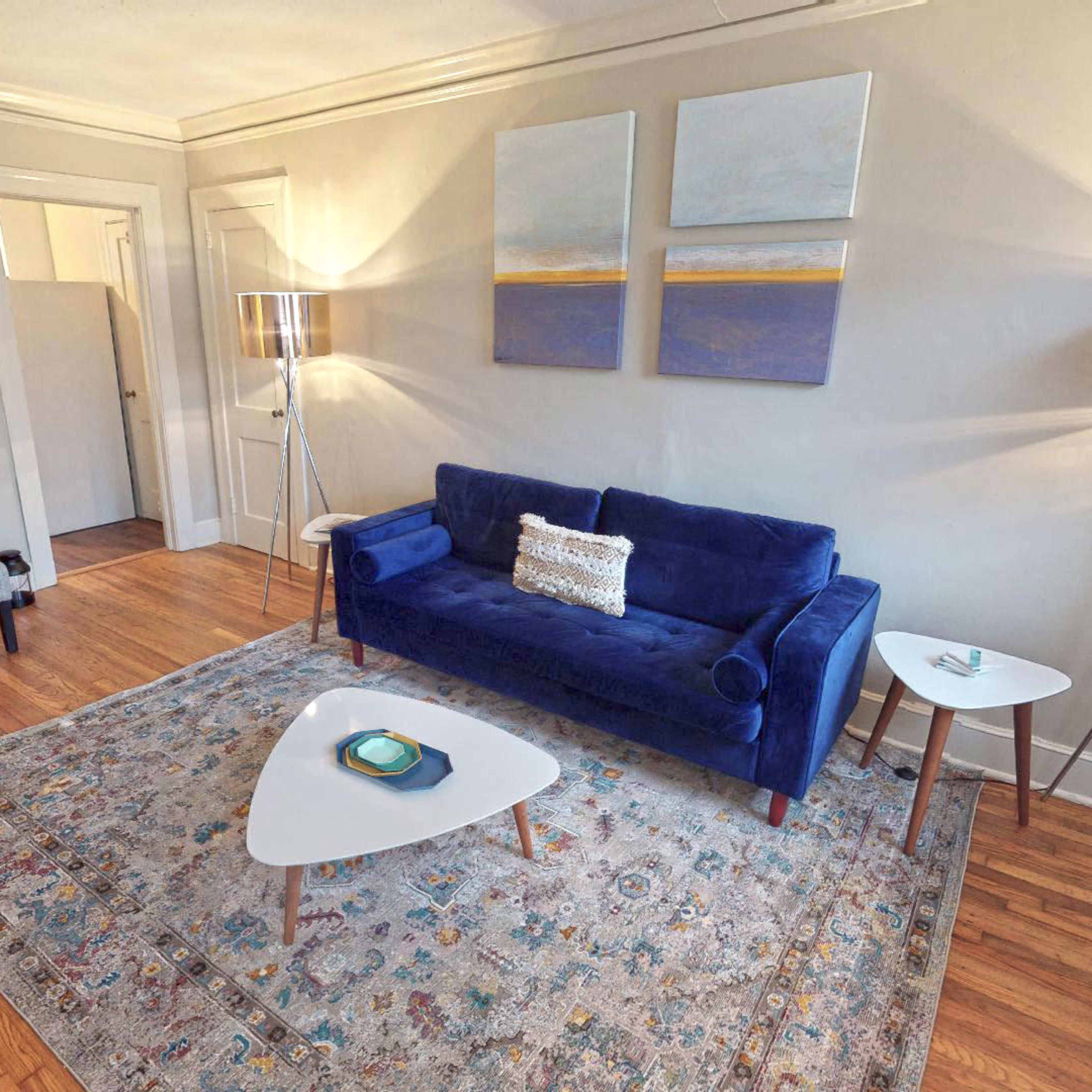 A living room featuring a blue sofa, two floor lamps, a small side table, and a patterned area rug on wooden flooring.