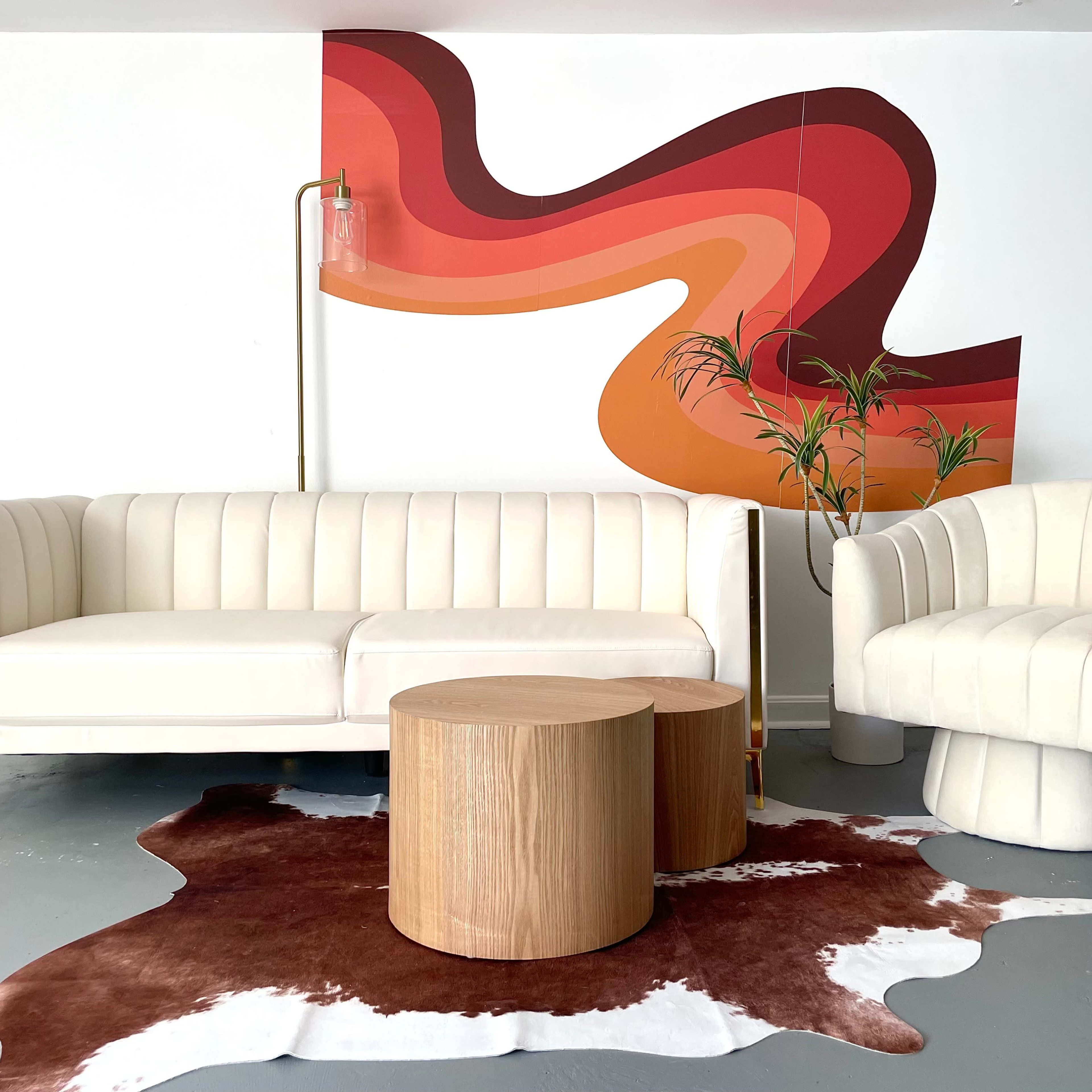 A modern living room setup with a white, ribbed sofa and armchair, a pair of wooden side tables, and a colorful abstract mural on the wall behind them.