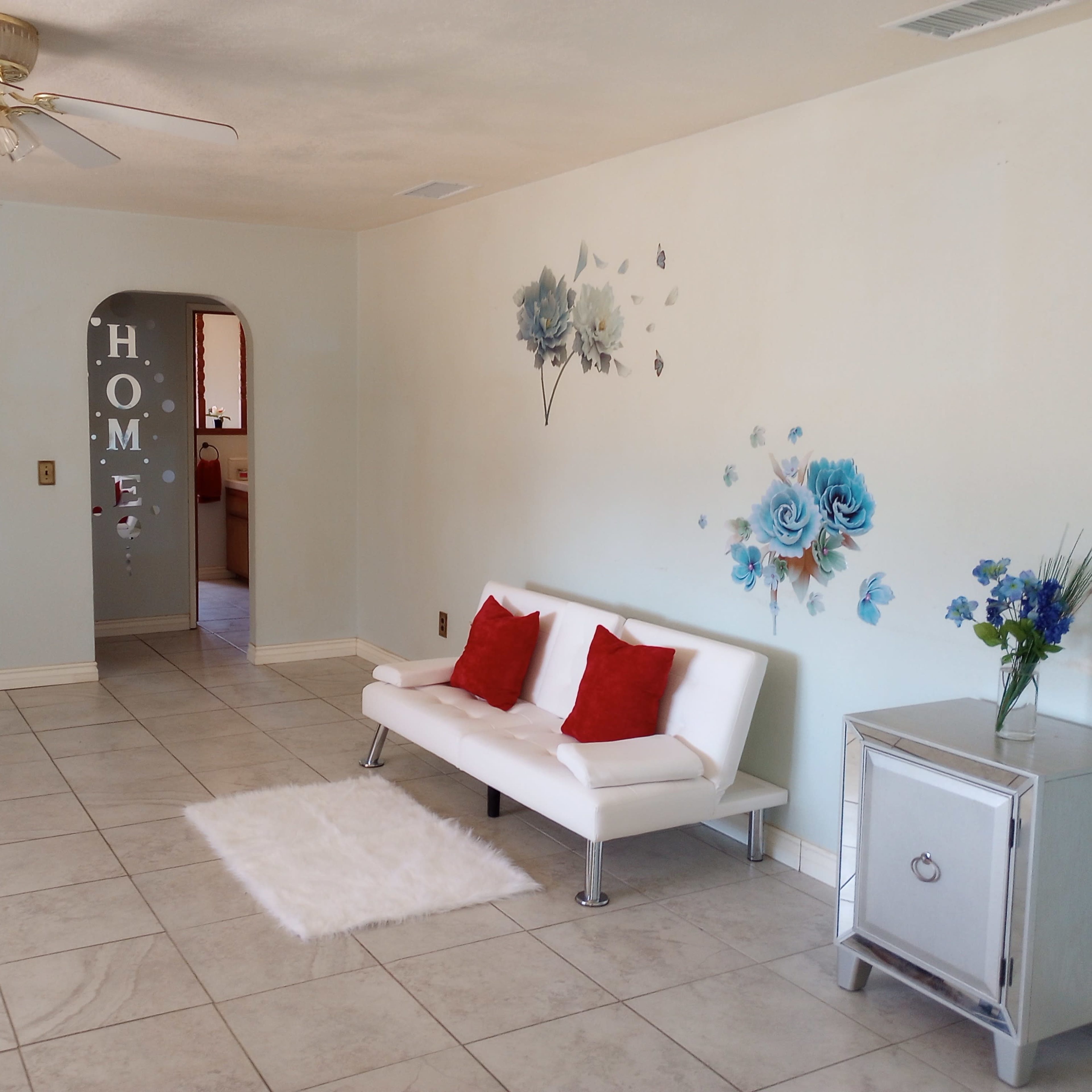 A living room with a white sofa adorned with red cushions, a small silver cabinet, and a floral wall decoration.