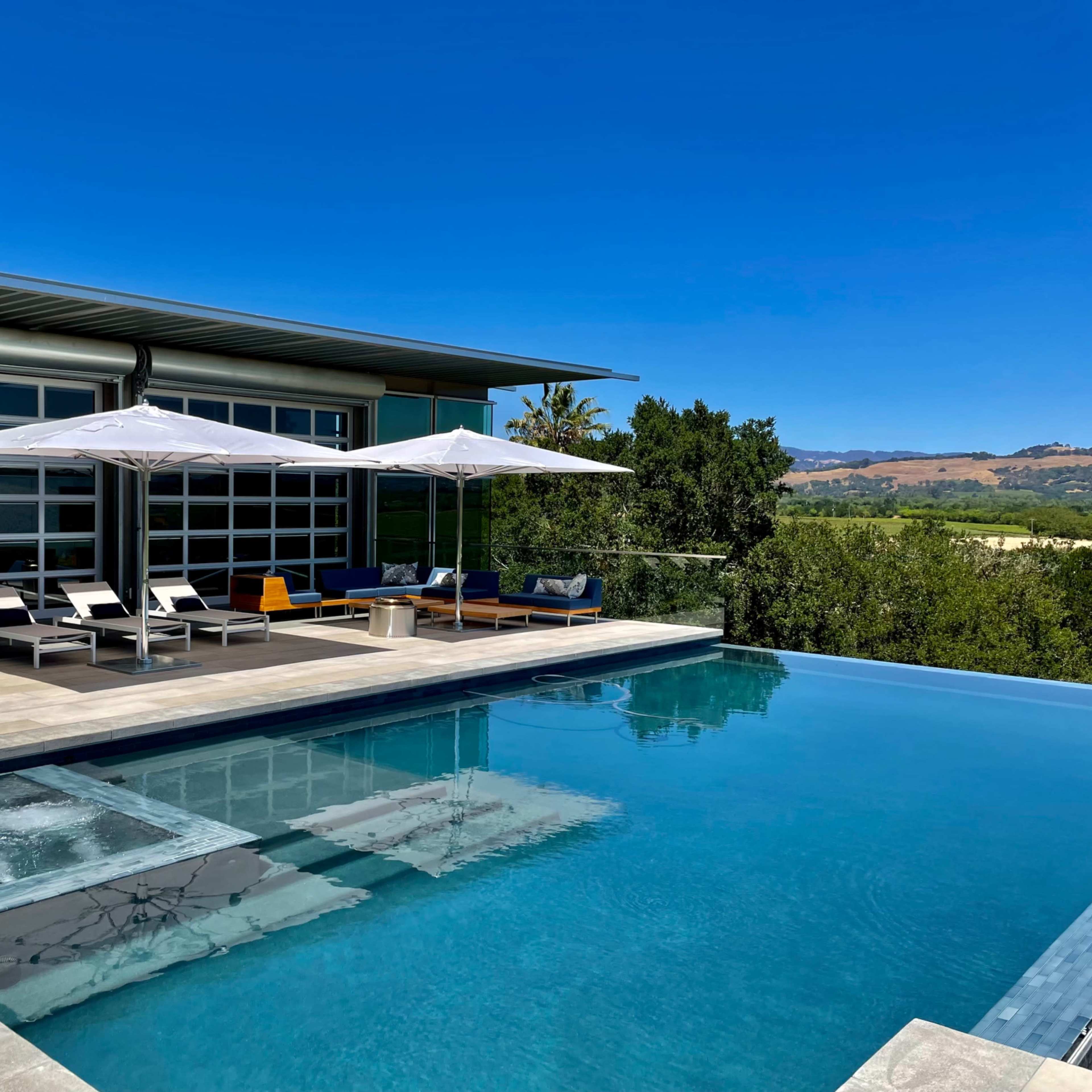A modern home with large windows overlooks a pool area featuring lounge chairs and sun umbrellas.