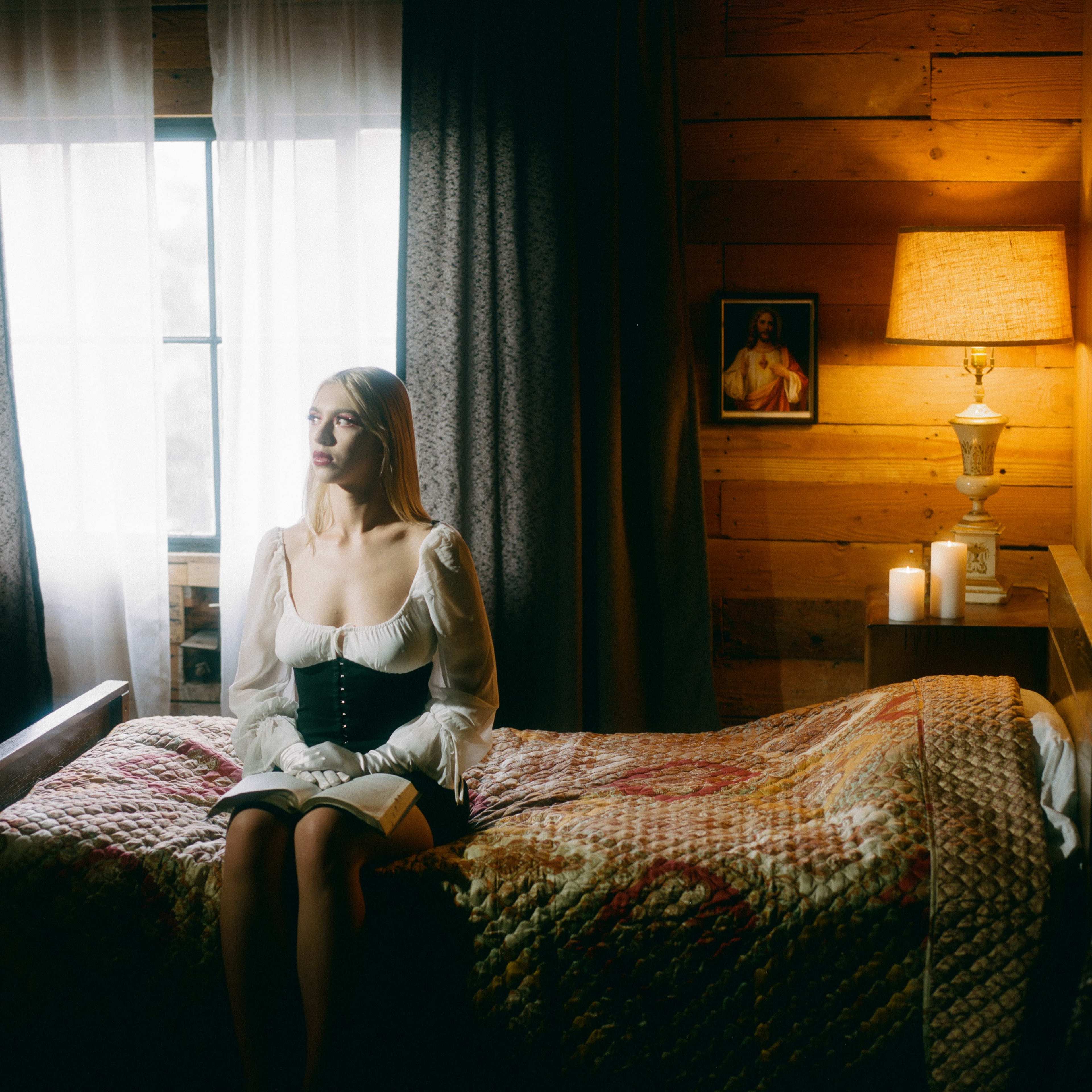 A woman sits on a bed in a room with wooden walls, reading a book beside a lamp and candles, with a window letting in natural light.