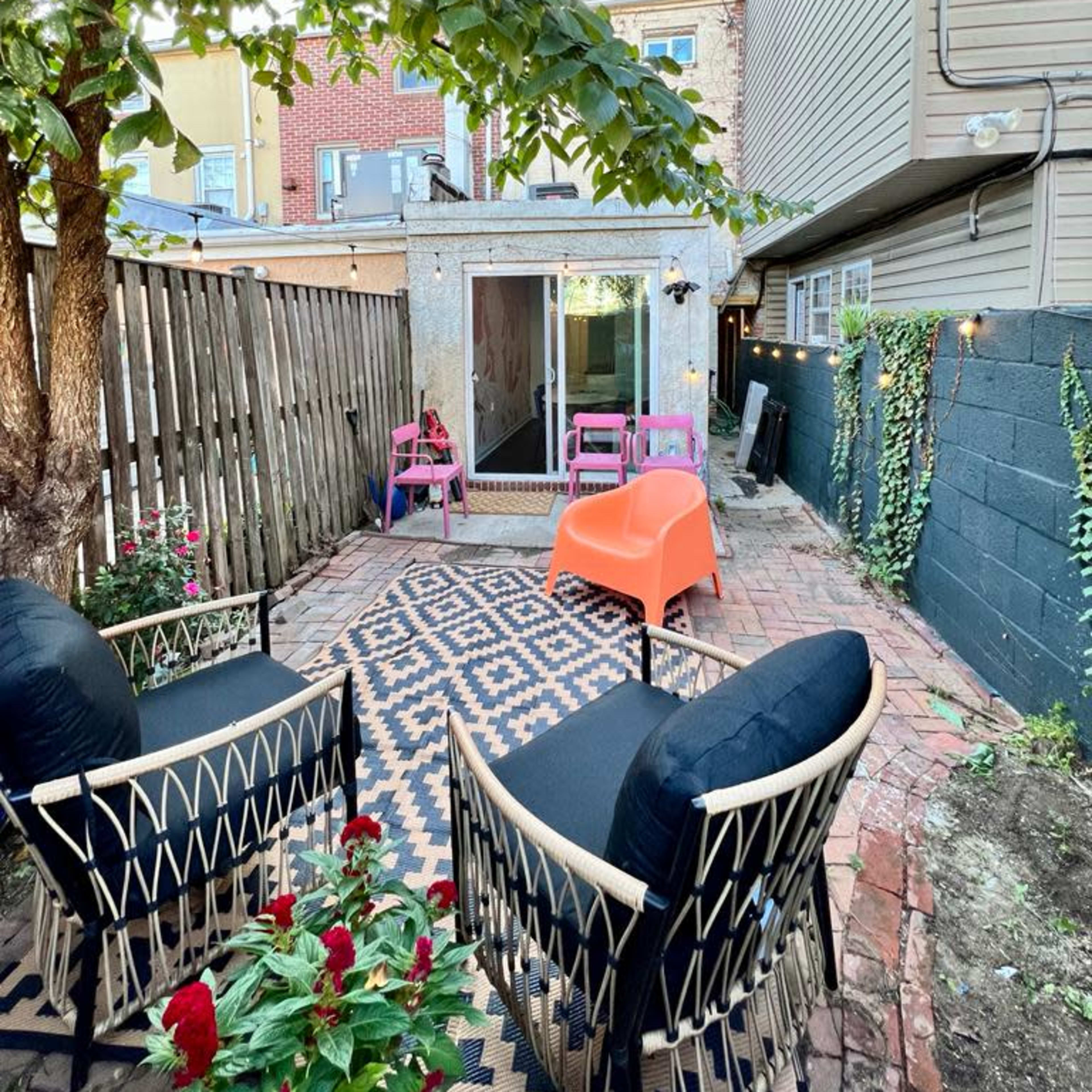 A small backyard features two black chairs and an orange chair arranged around a patterned rug, with a shed in the background surrounded by greenery.