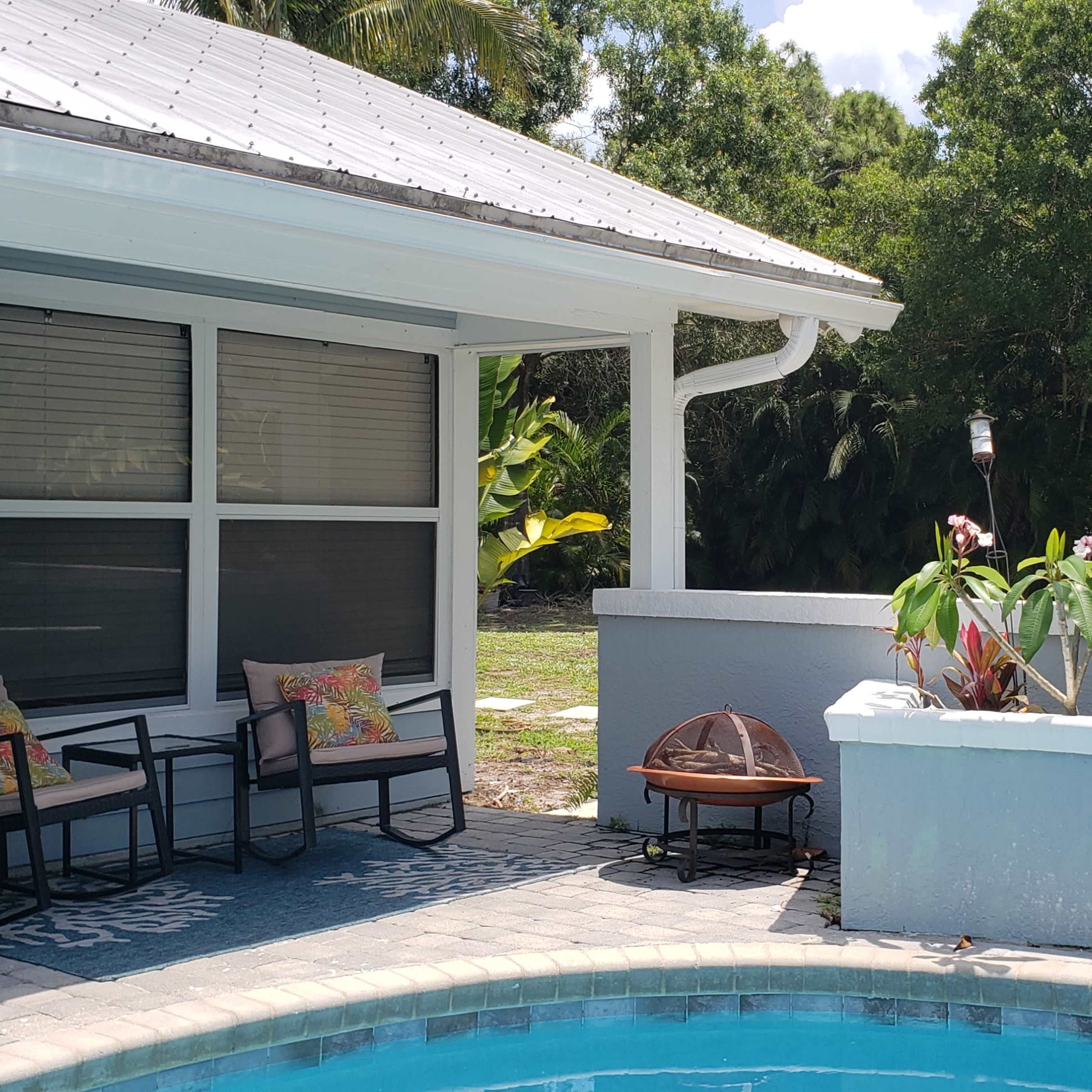 A swimming pool with a patio area featuring seating and potted plants, set against a backdrop of lush greenery.