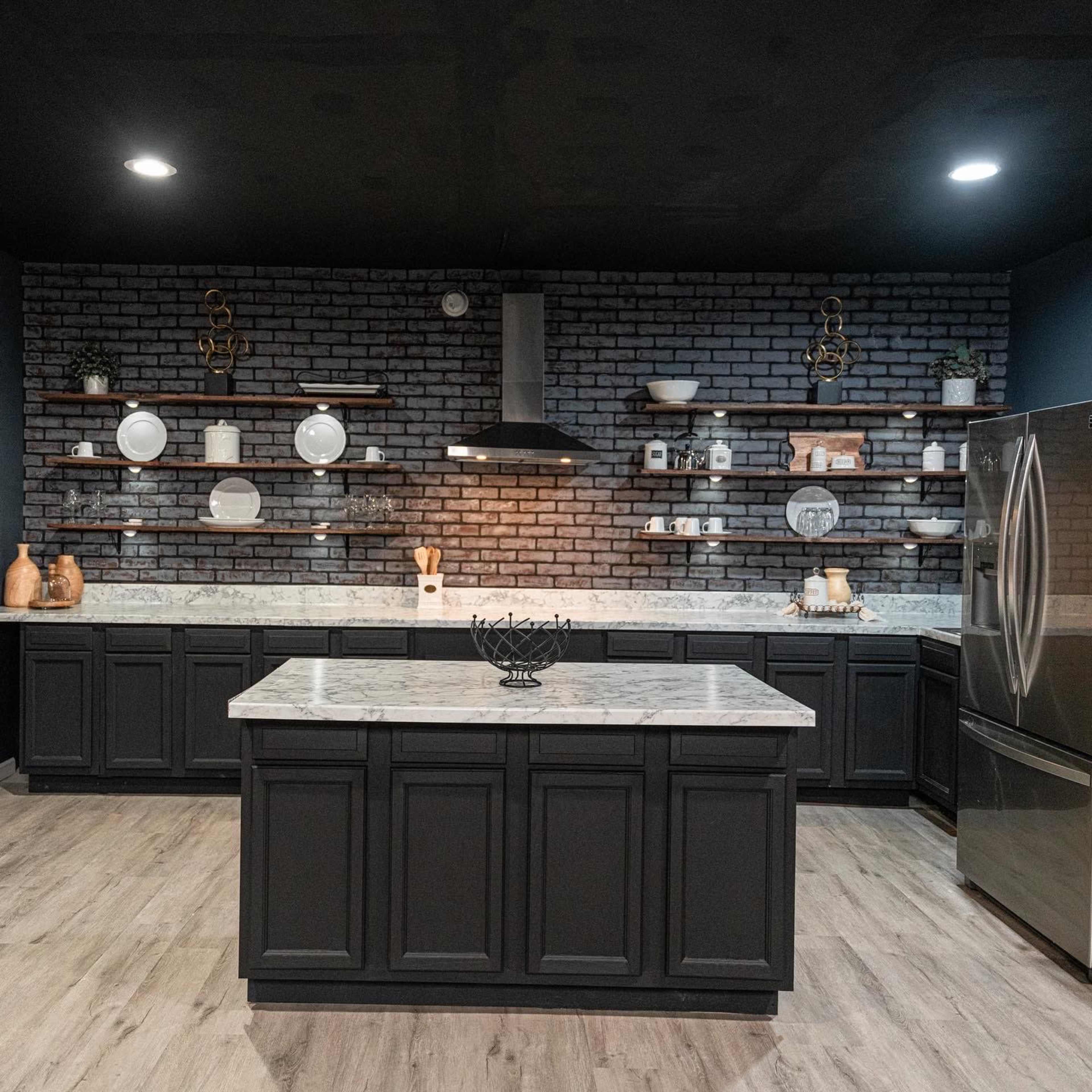 The kitchen features dark cabinetry, a marble countertop island, and a brick accent wall with wooden shelves displaying dinnerware.