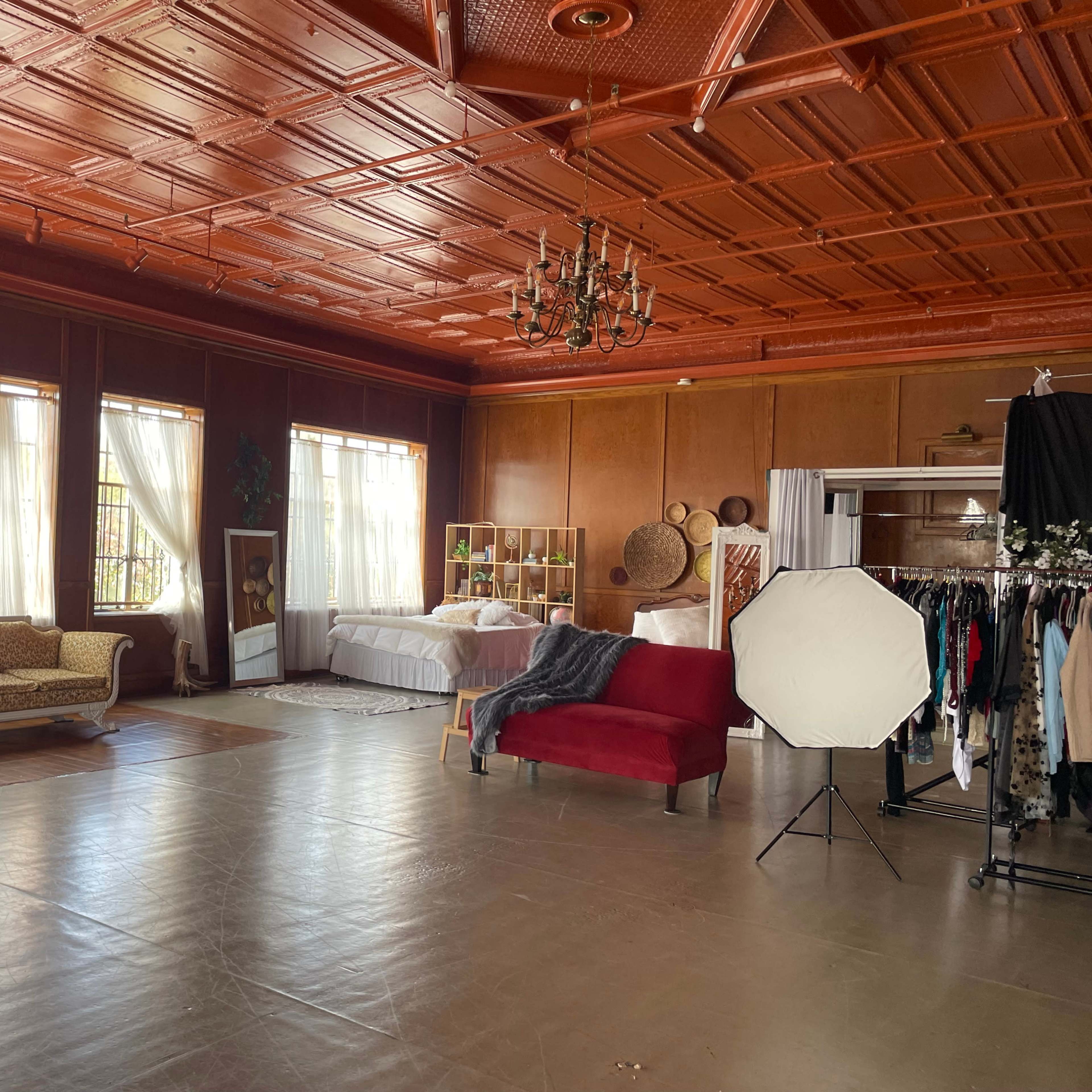 A spacious room with wooden ceiling, large windows allowing natural light, and a mix of furniture including a yellow sofa and a red armchair, along with clothing racks and a large photography reflector.