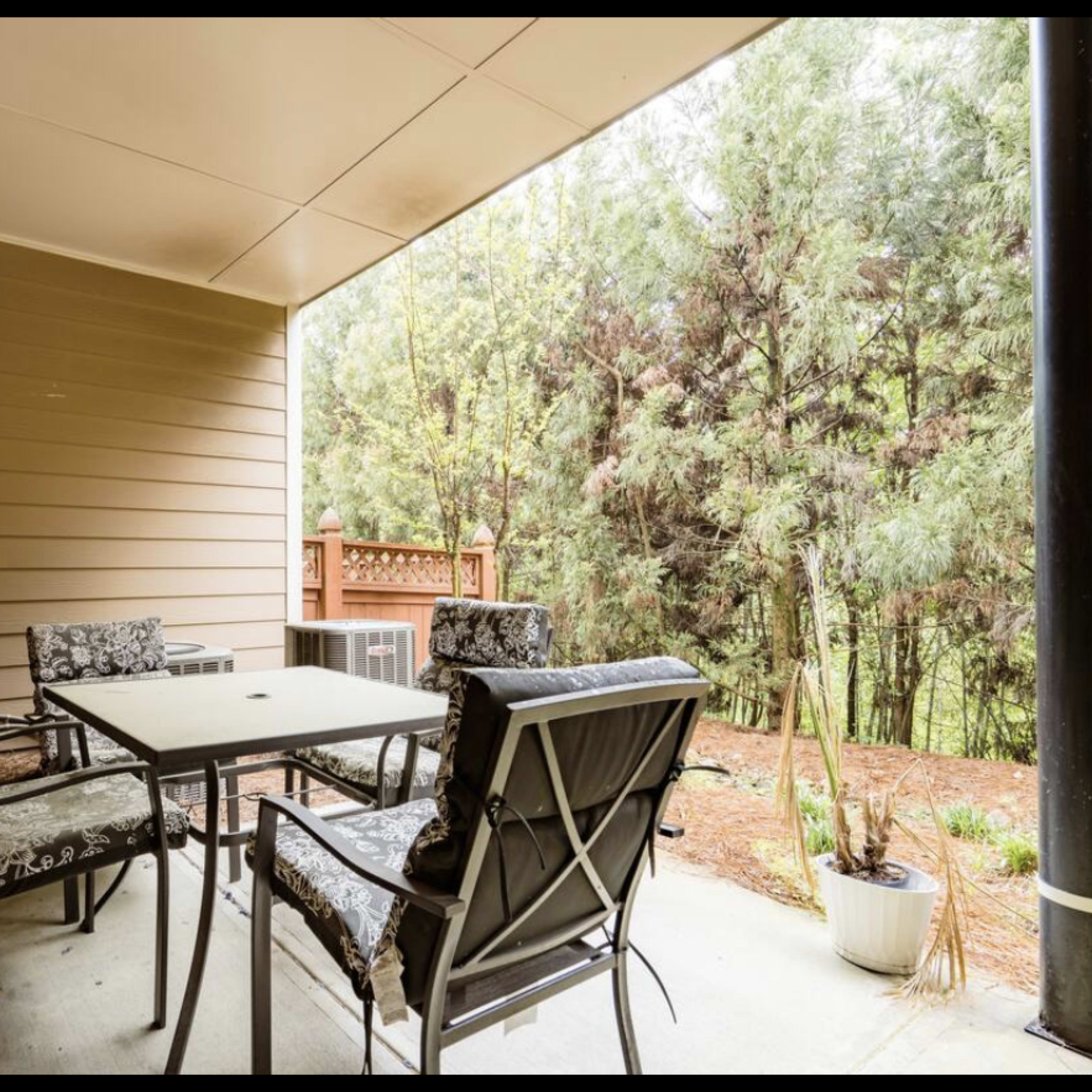 A covered patio area with a table and four chairs surrounded by greenery.