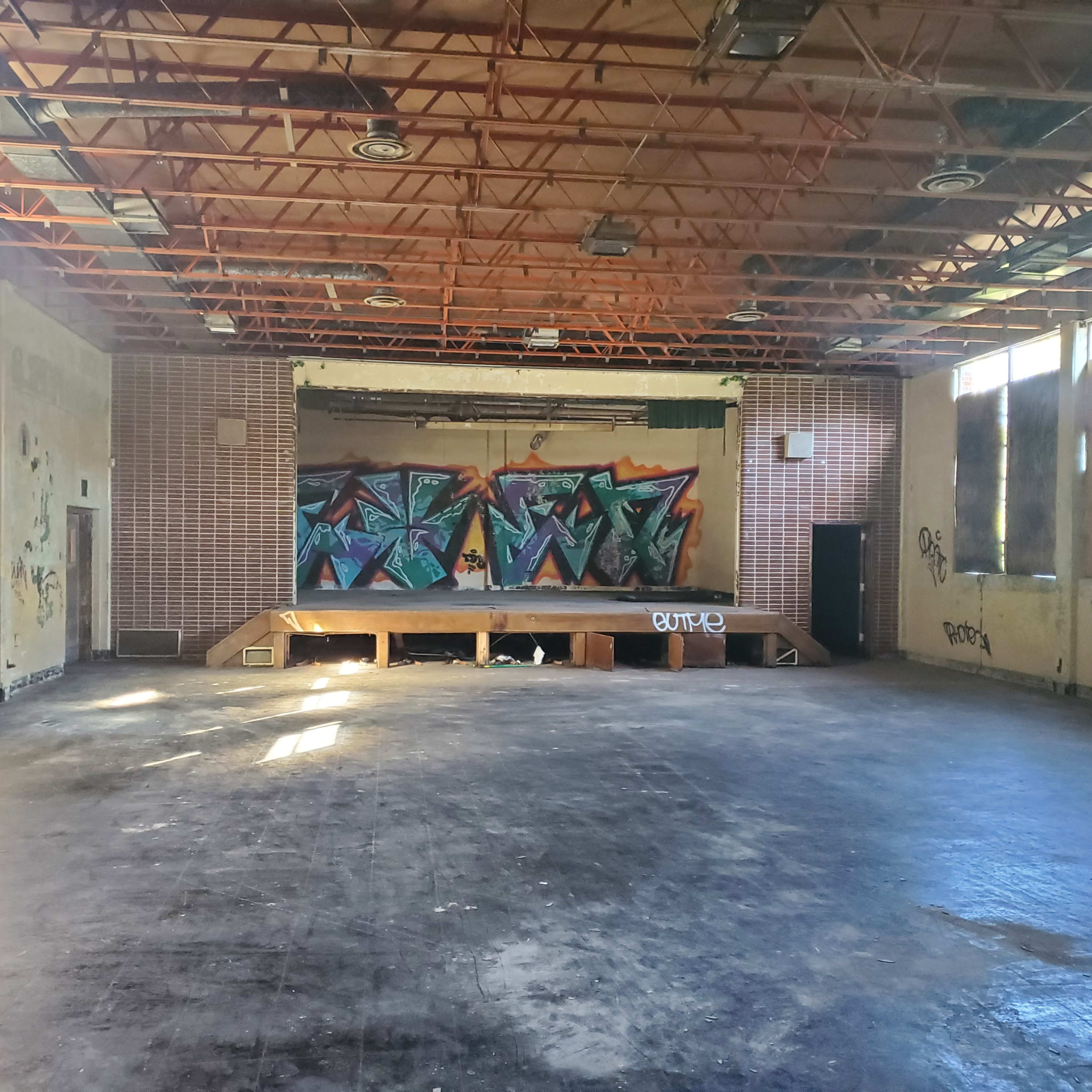 An empty, dilapidated room with a wooden stage and colorful graffiti on the back wall.