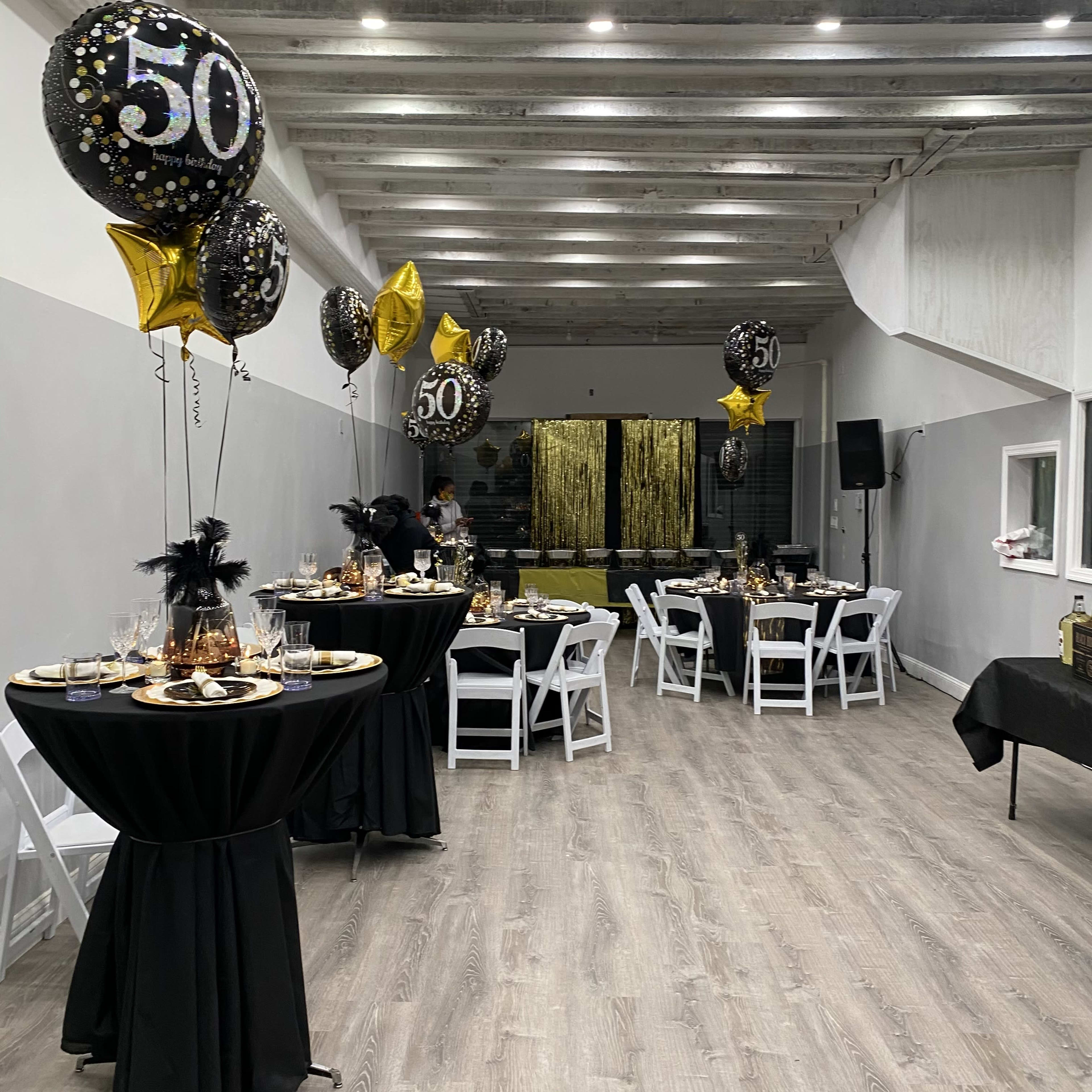 A decorated event space featuring black and gold balloons for a 50th birthday celebration, with elegantly set tables and a shimmering backdrop.