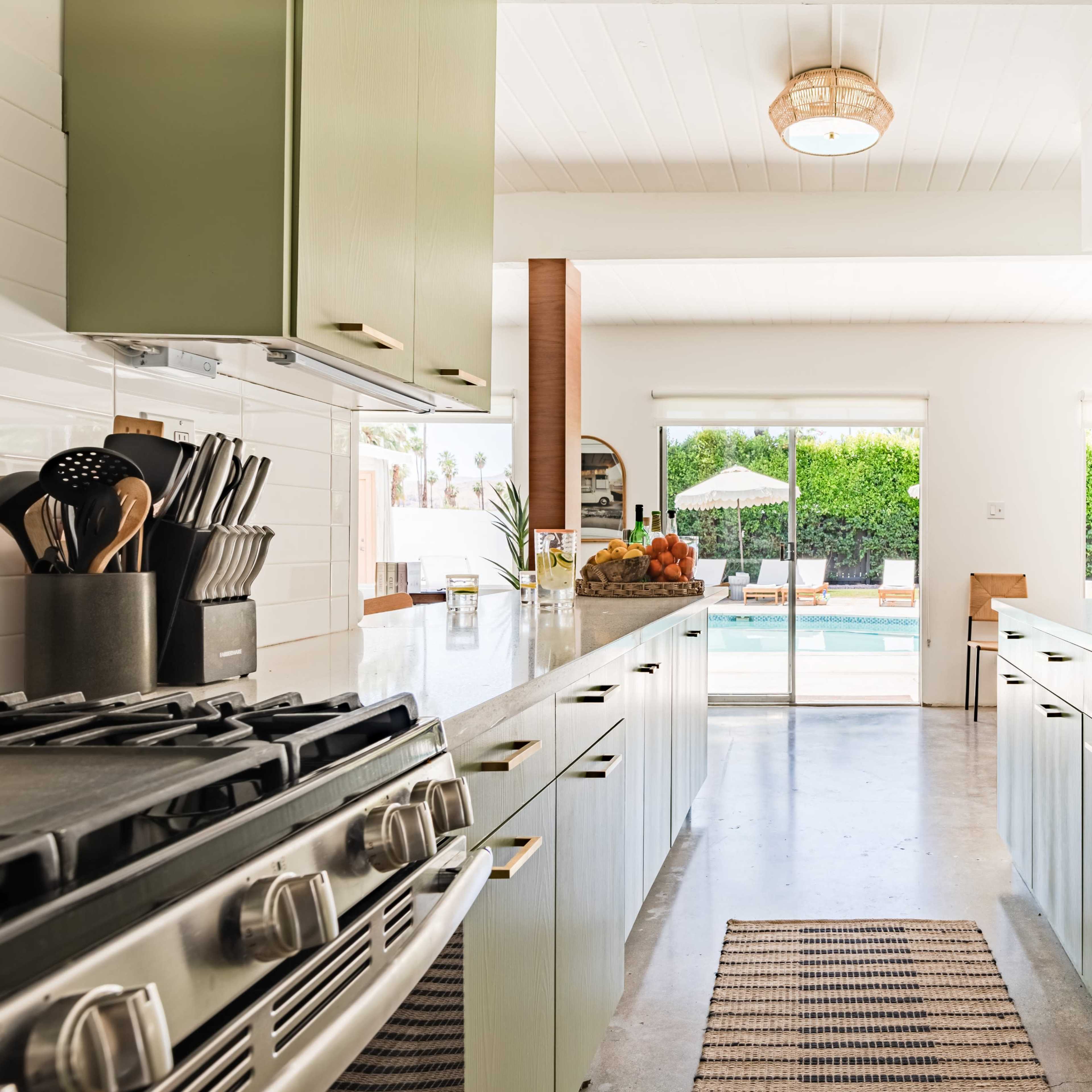 A modern kitchen features green cabinets, a large island with a gas stove, and a view of a pool area through sliding glass doors.