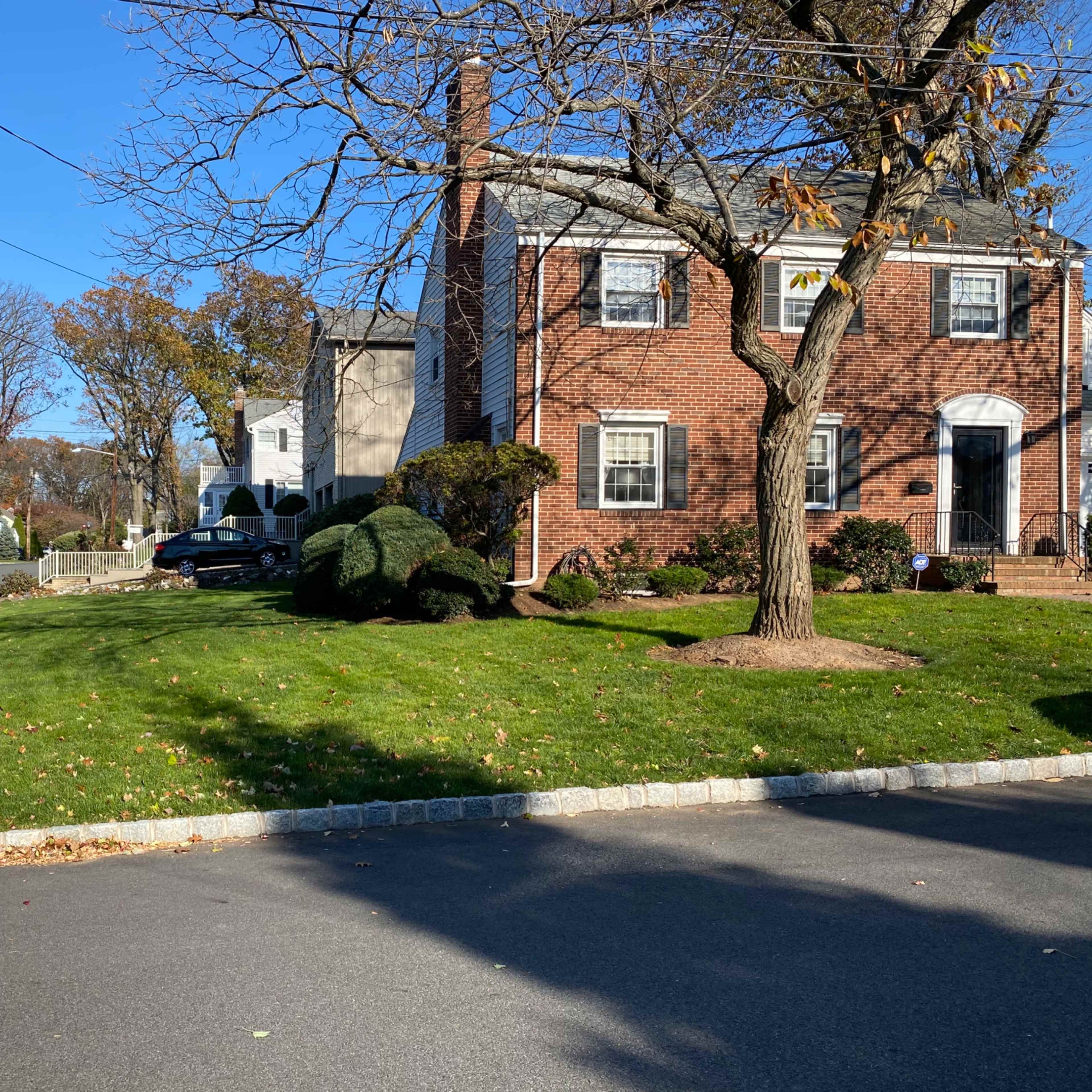A brick house with white trim is situated on a grassy corner lot, surrounded by neatly trimmed bushes and trees, with a clear blue sky overhead.
