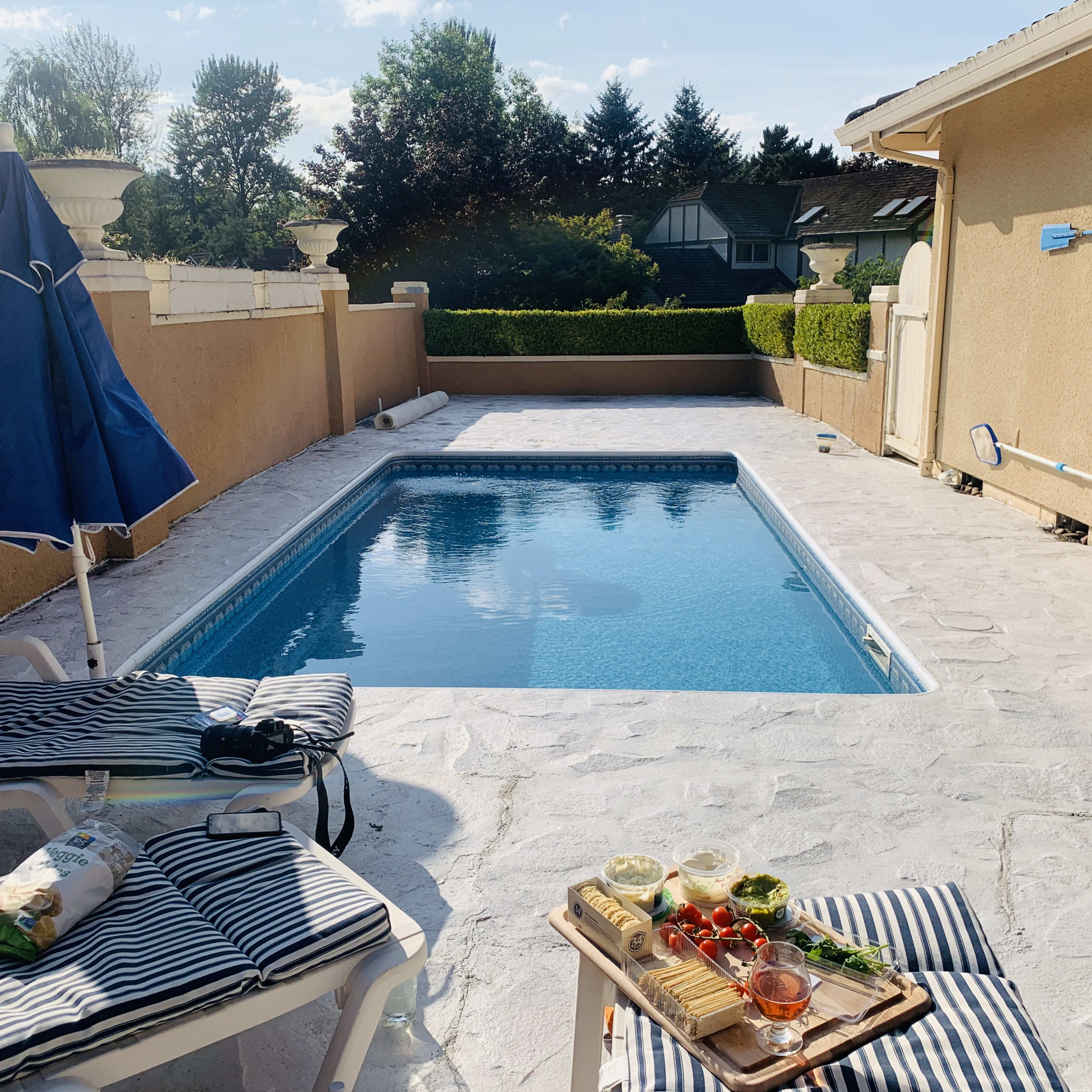 A backyard pool area features a blue pool surrounded by lounge chairs and a table with various food items.