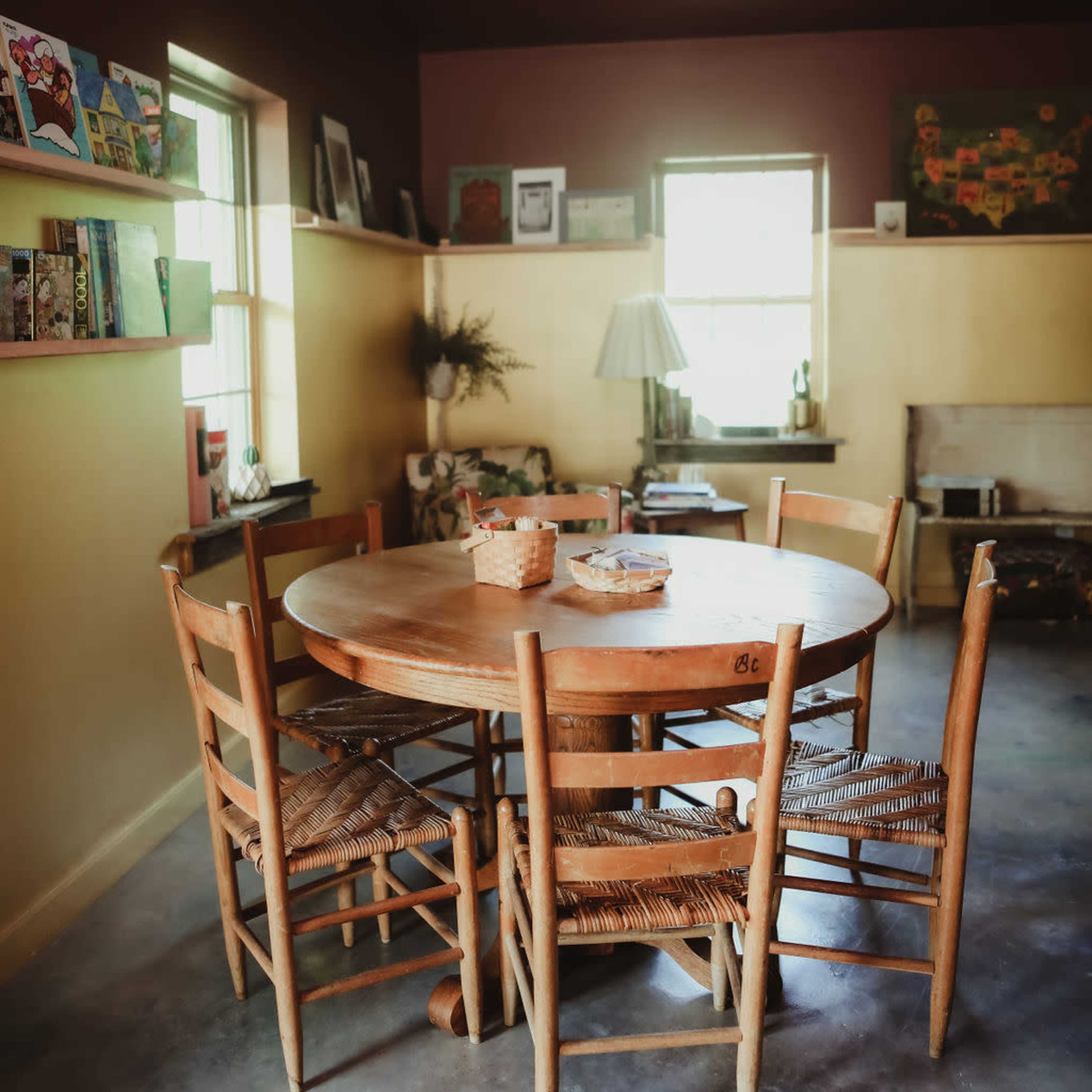 A round wooden dining table surrounded by six chairs is set in a warmly lit room with artwork on the walls.