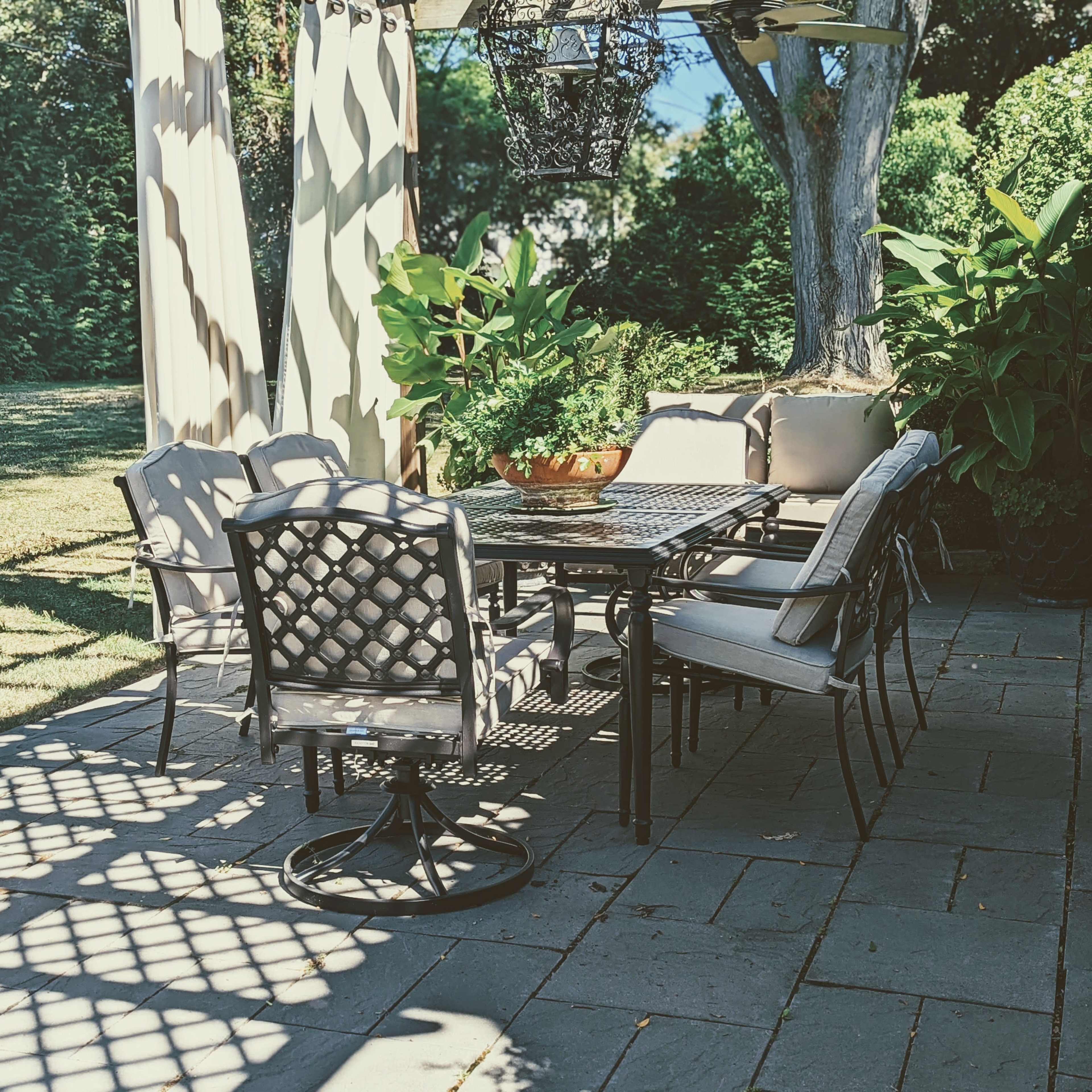 A patio seating area features a metal dining table surrounded by six chairs, shaded by a pergola and illuminated by hanging lanterns, with greenery in the background.