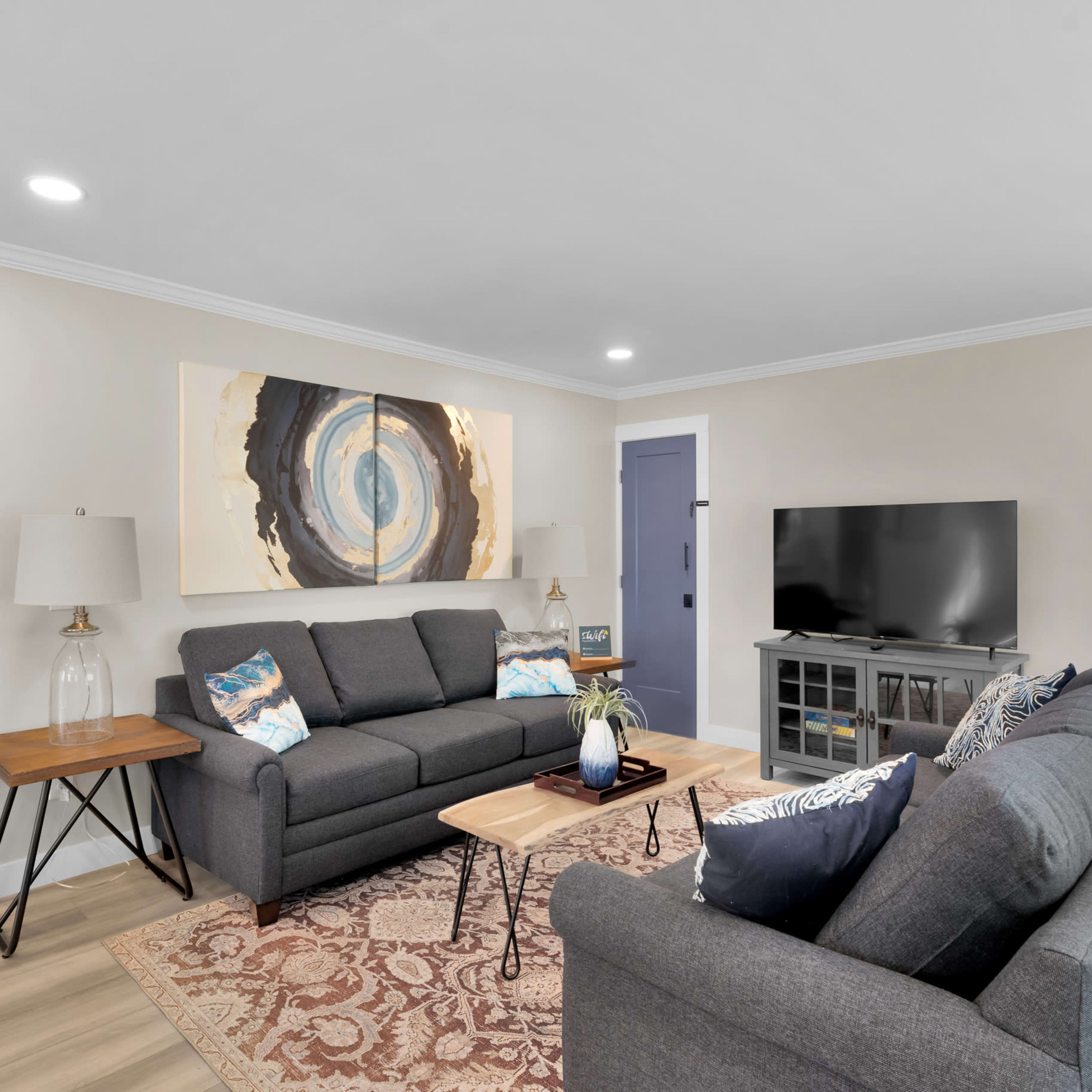 A living room features two gray sofas, a wooden coffee table, a television on a stand, and artwork on the wall.