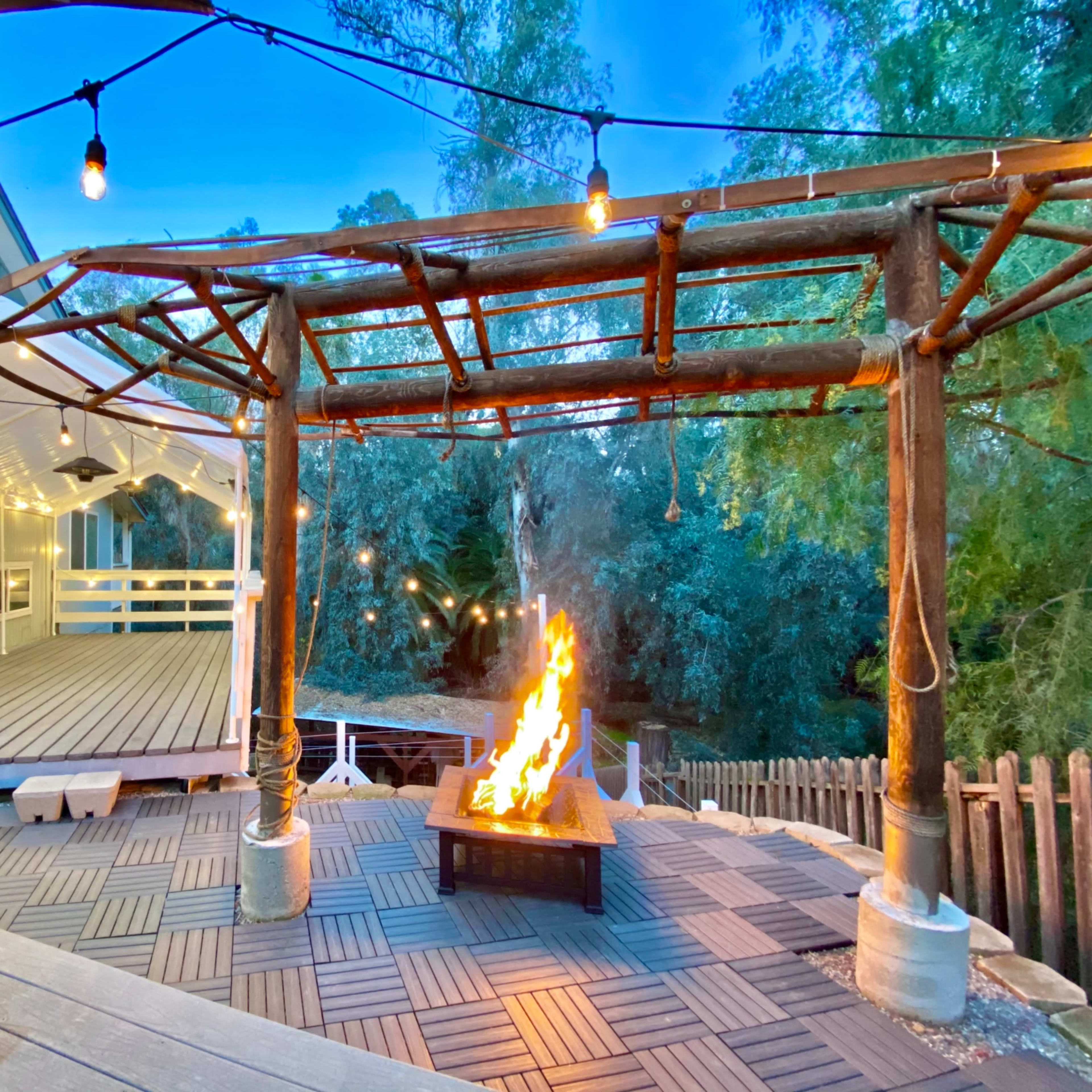 A wooden deck features a fire pit surrounded by string lights and a pergola, with greenery visible in the background.