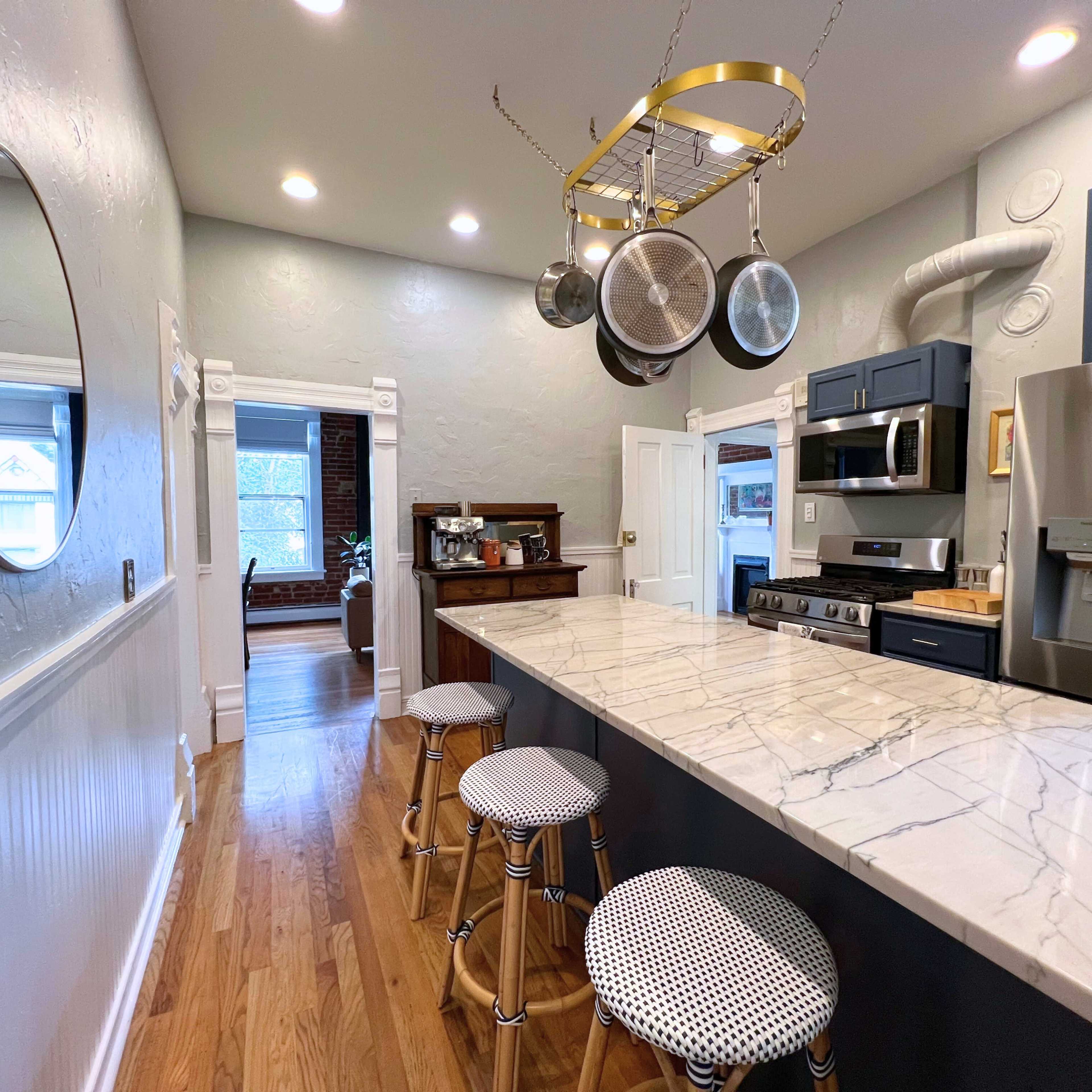 A modern kitchen features a marble countertop, blue cabinets, and a circular mirror on the wall, with three stools arranged at the counter.