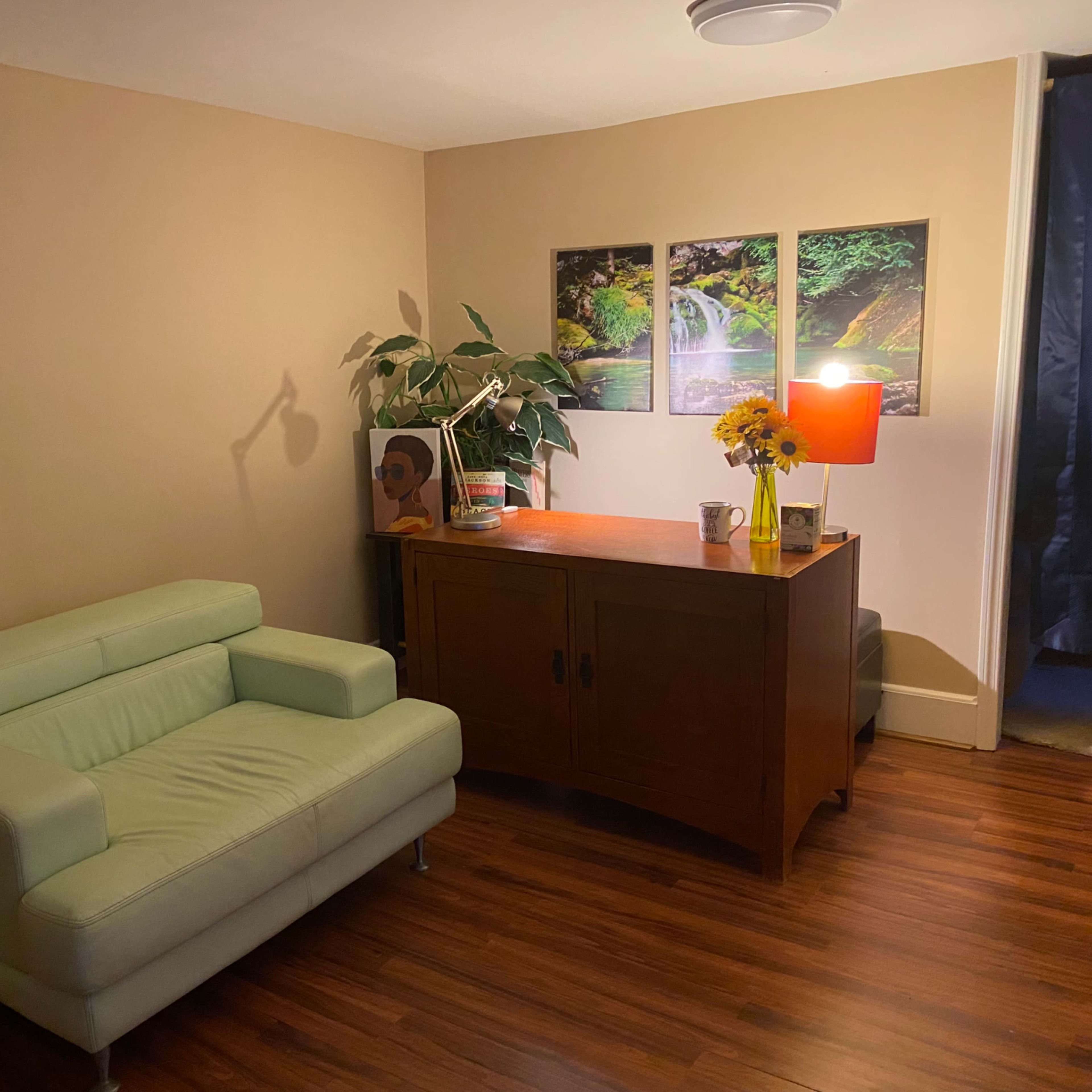 A small living room with a light green sofa, a wooden side table, a lamp, a vase of flowers, and paintings of waterfalls on the wall.