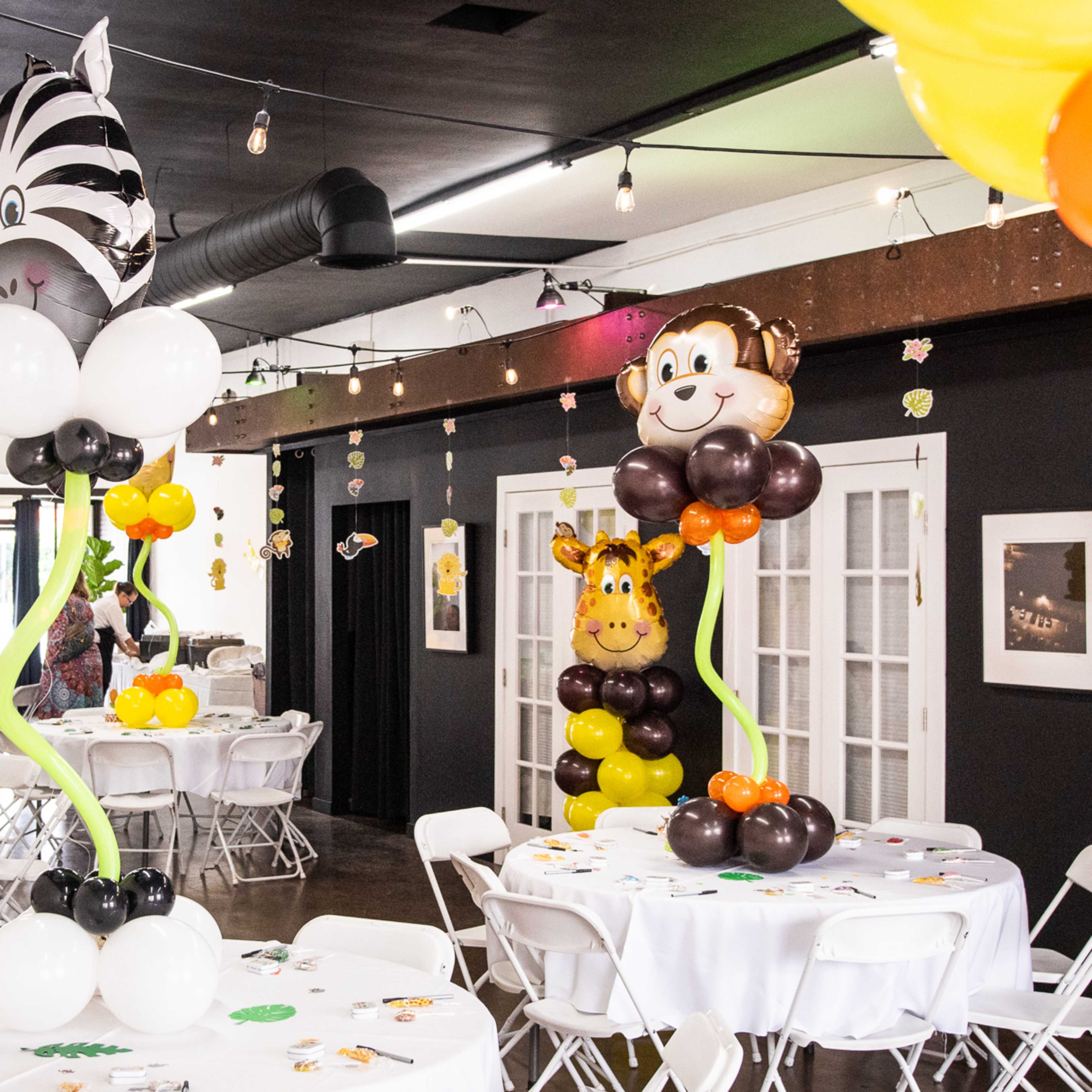 A decorated event space with tables covered in white cloth, filled with colorful balloon centerpieces in the shapes of animals, and festive balloons hanging from the ceiling.