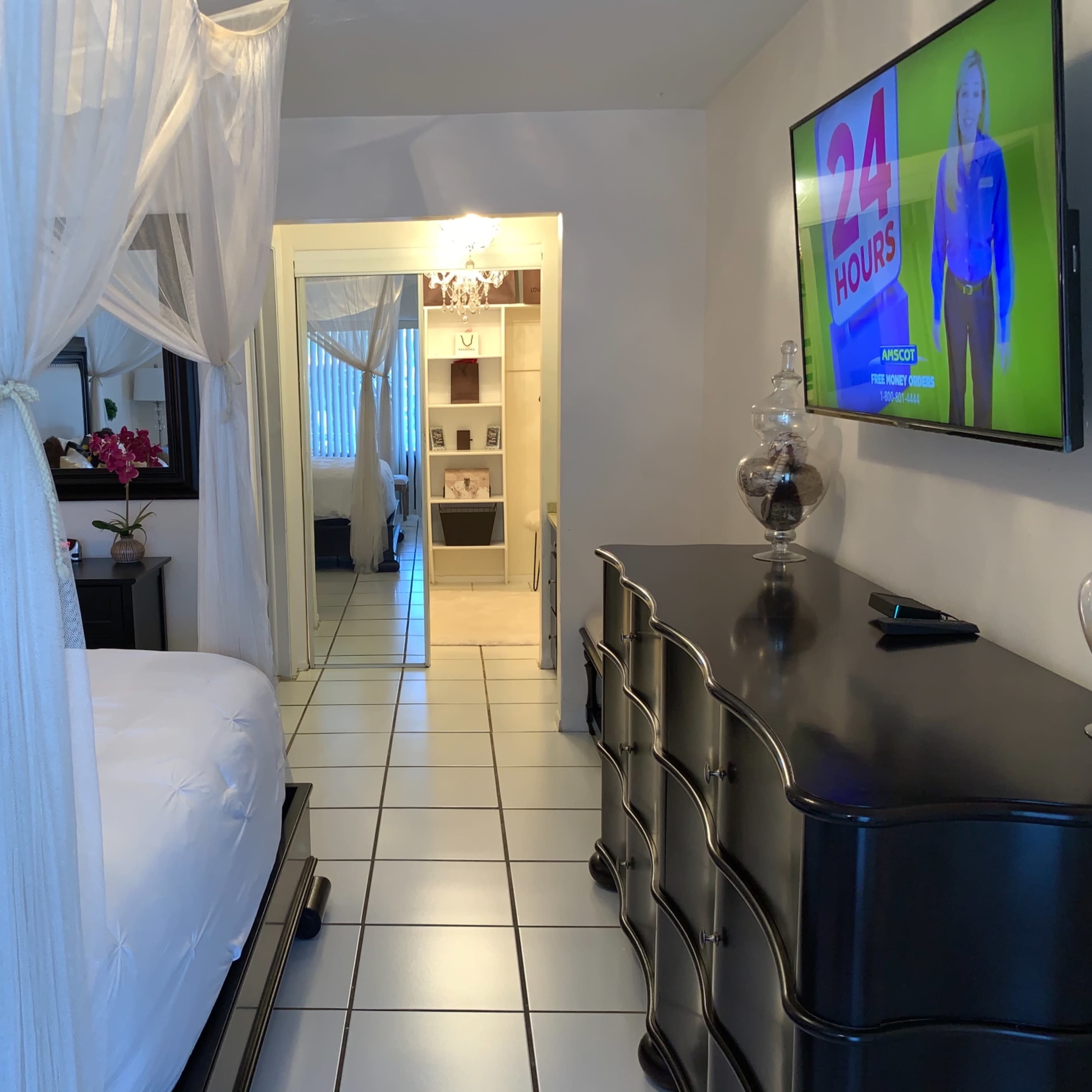 A bedroom with a four-poster bed, a dresser, and a wall-mounted television, leading into a mirrored hallway.